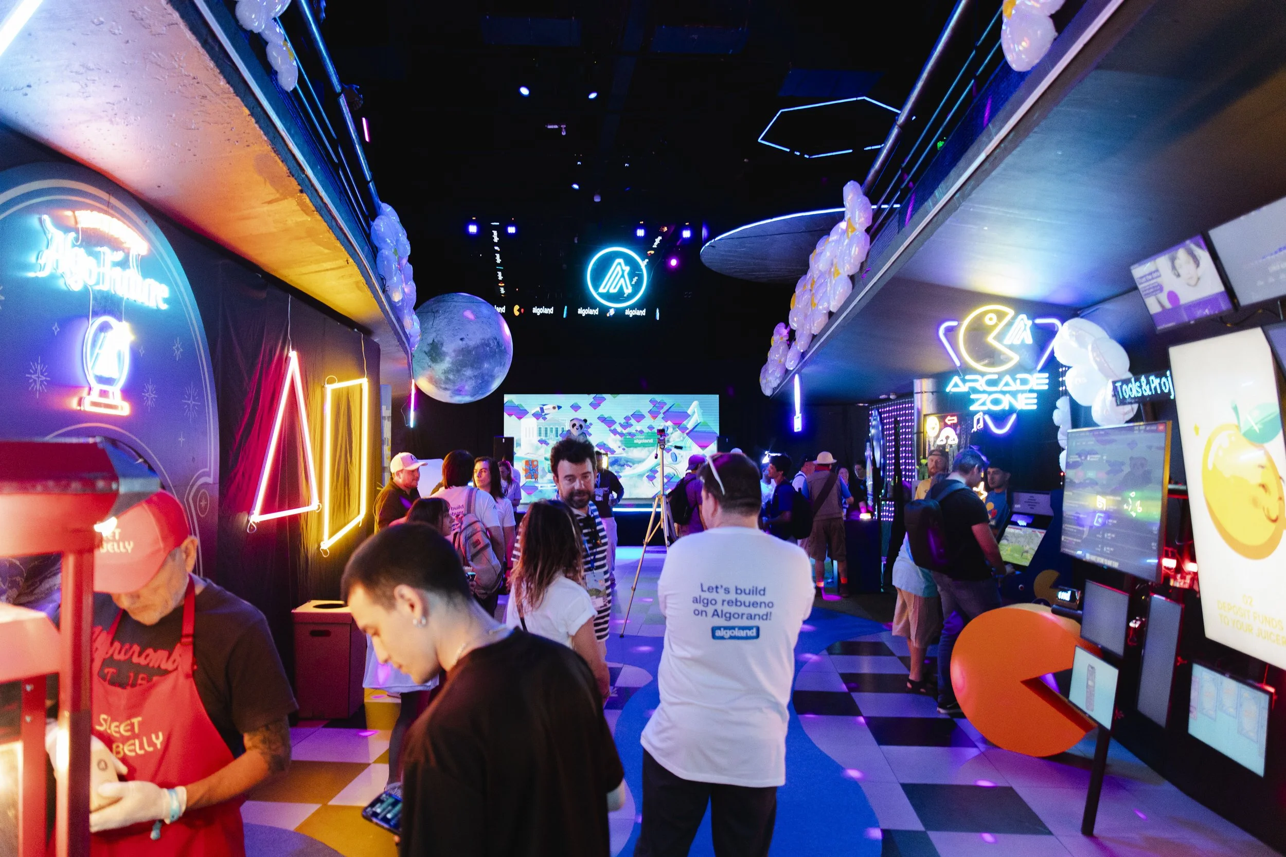 People playing arcade games and socializing in a brightly lit arcade zone with neon signs, colorful lighting, and digital screens.