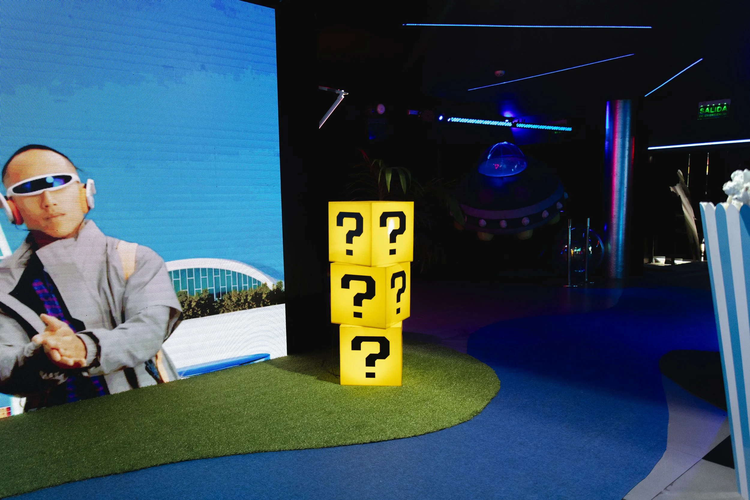 A display with four illuminated yellow boxes, each with a black question mark on it, stacked in a 2x2 formation on a small patch of artificial grass.