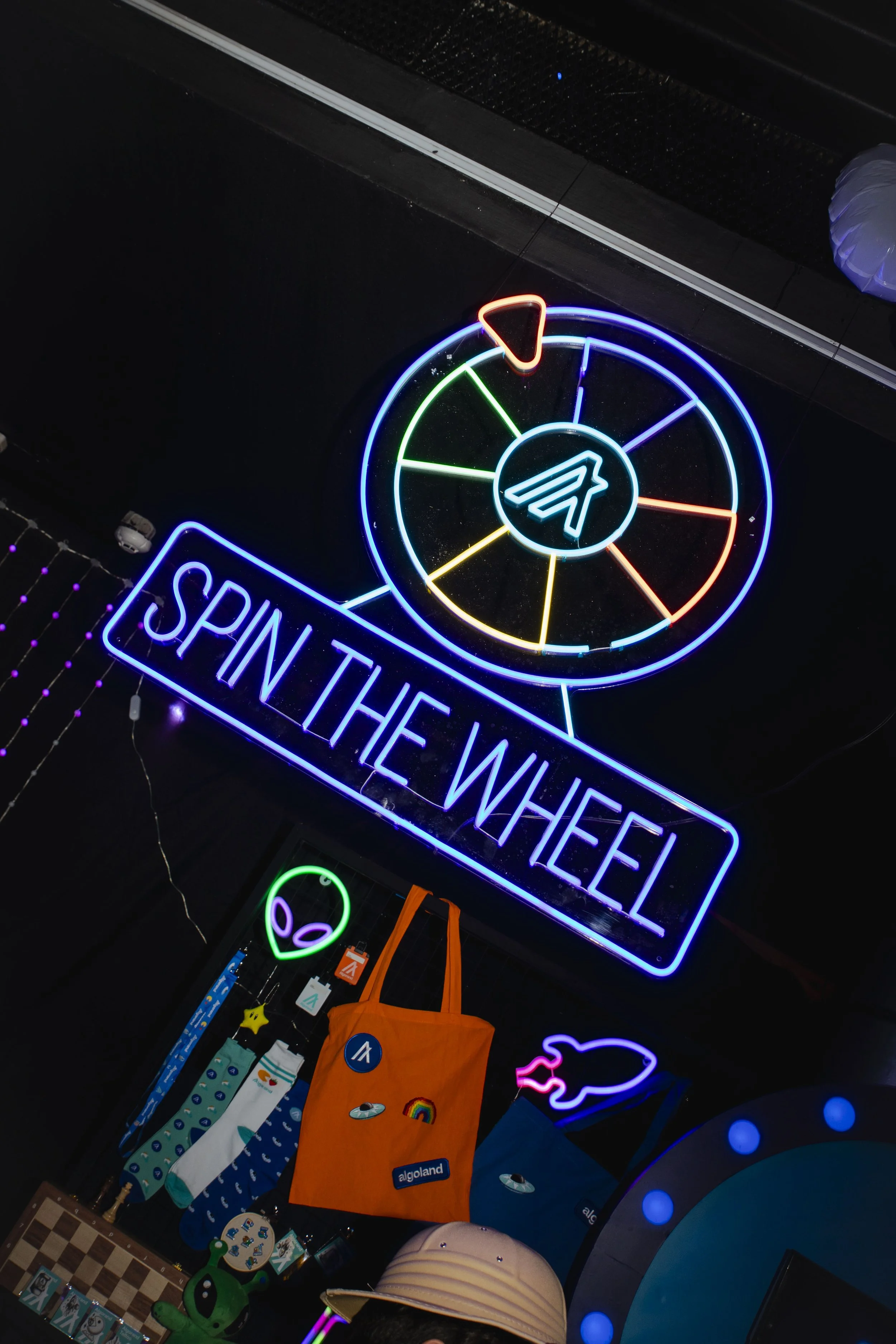 Neon sign that says "Spin the Wheel" with a colorful spinning wheel above it, and various decorations including alien plush toys, socks, a tote bag, and other items are visible in the background.