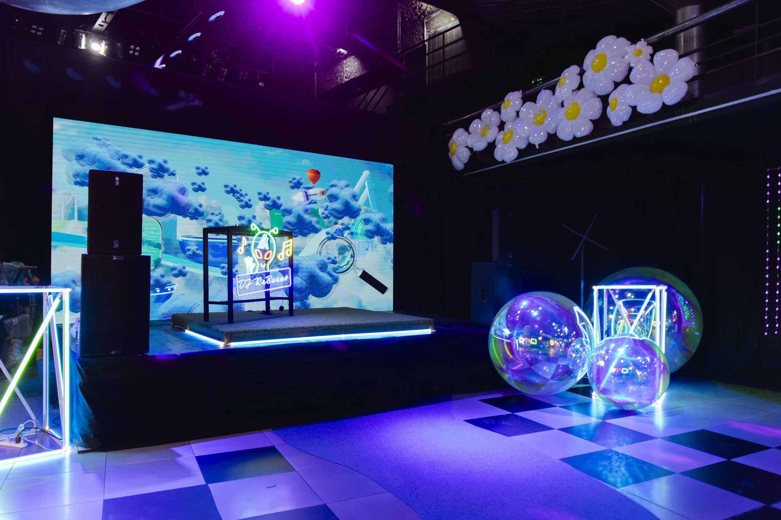 A decorated party stage with neon lights and a background screen displaying colorful clouds and a hot air balloon. There are large inflatable transparent spheres with lights, a floral balloon arrangement hanging from the ceiling, and a black and whit