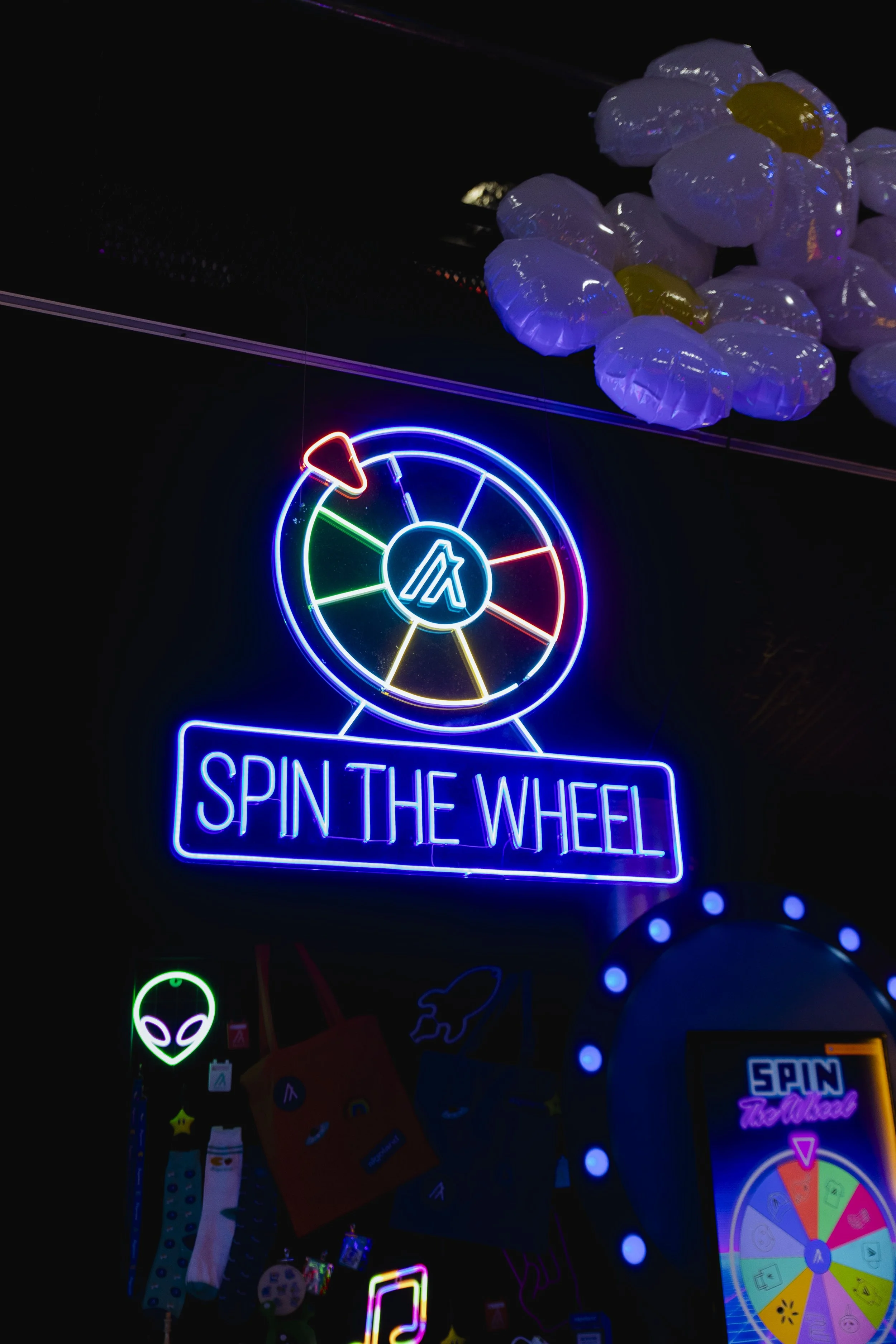 Neon sign with a spinning wheel and the phrase 'Spin the Wheel' in colorful lights, with plush flower decorations on the ceiling.