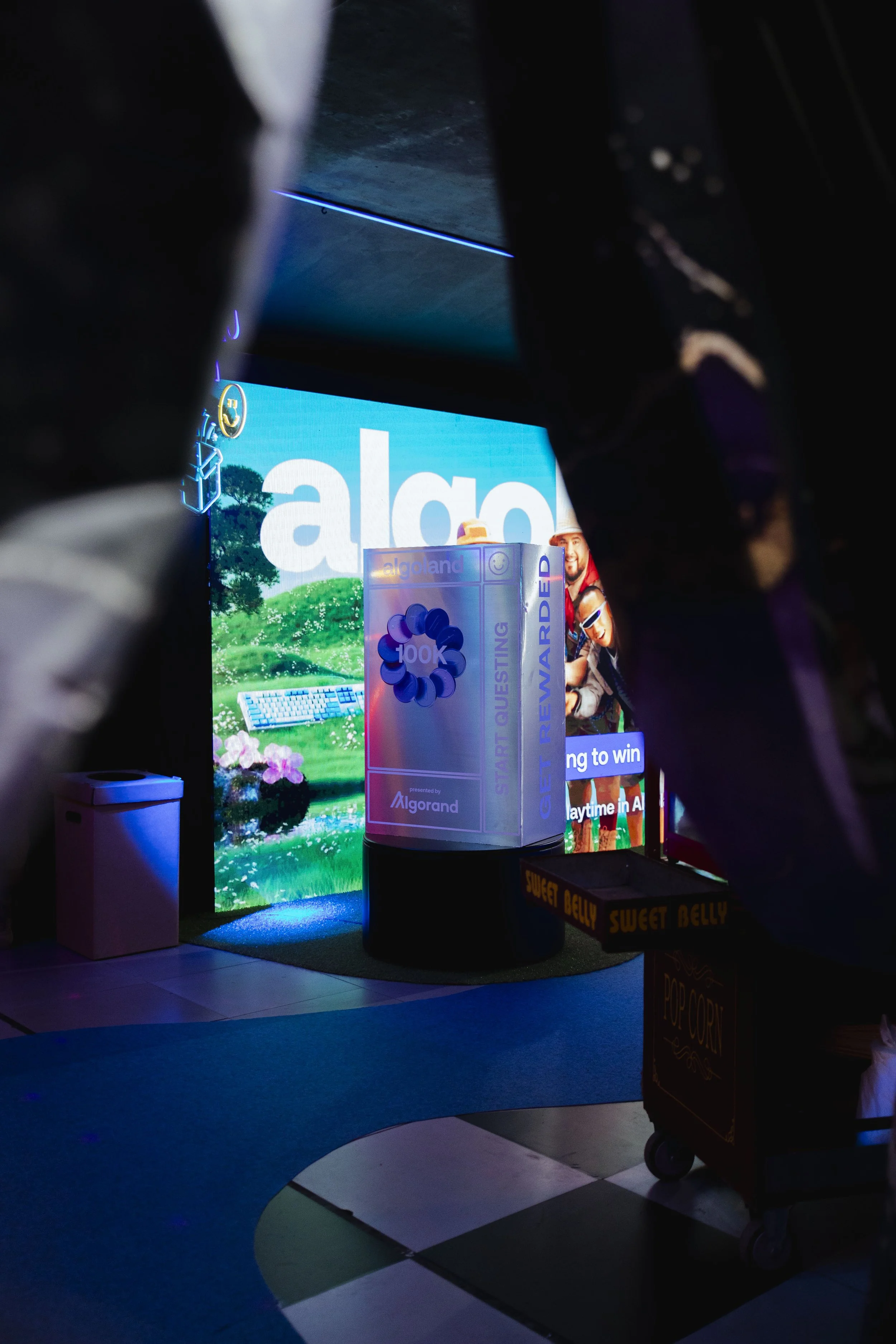 A digital display and a large prize box with Algorand branding seen through an opening, with a small white trash bin and a popcorn cart labeled 'Sweet Belly' in the scene, with a dark surrounding area.