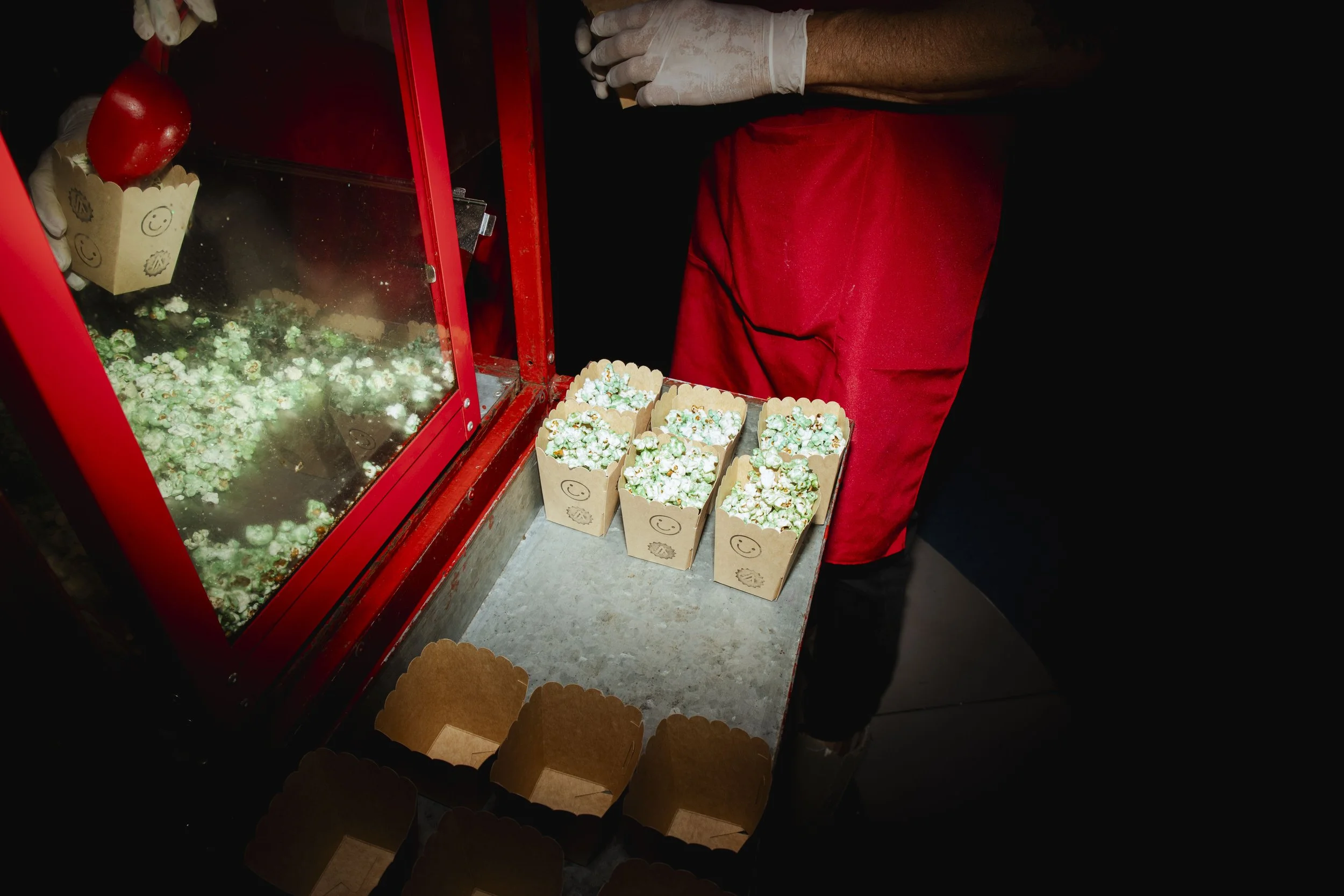 A person with white gloves in a red apron making popcorn at a popcorn machine with green chocolates on top