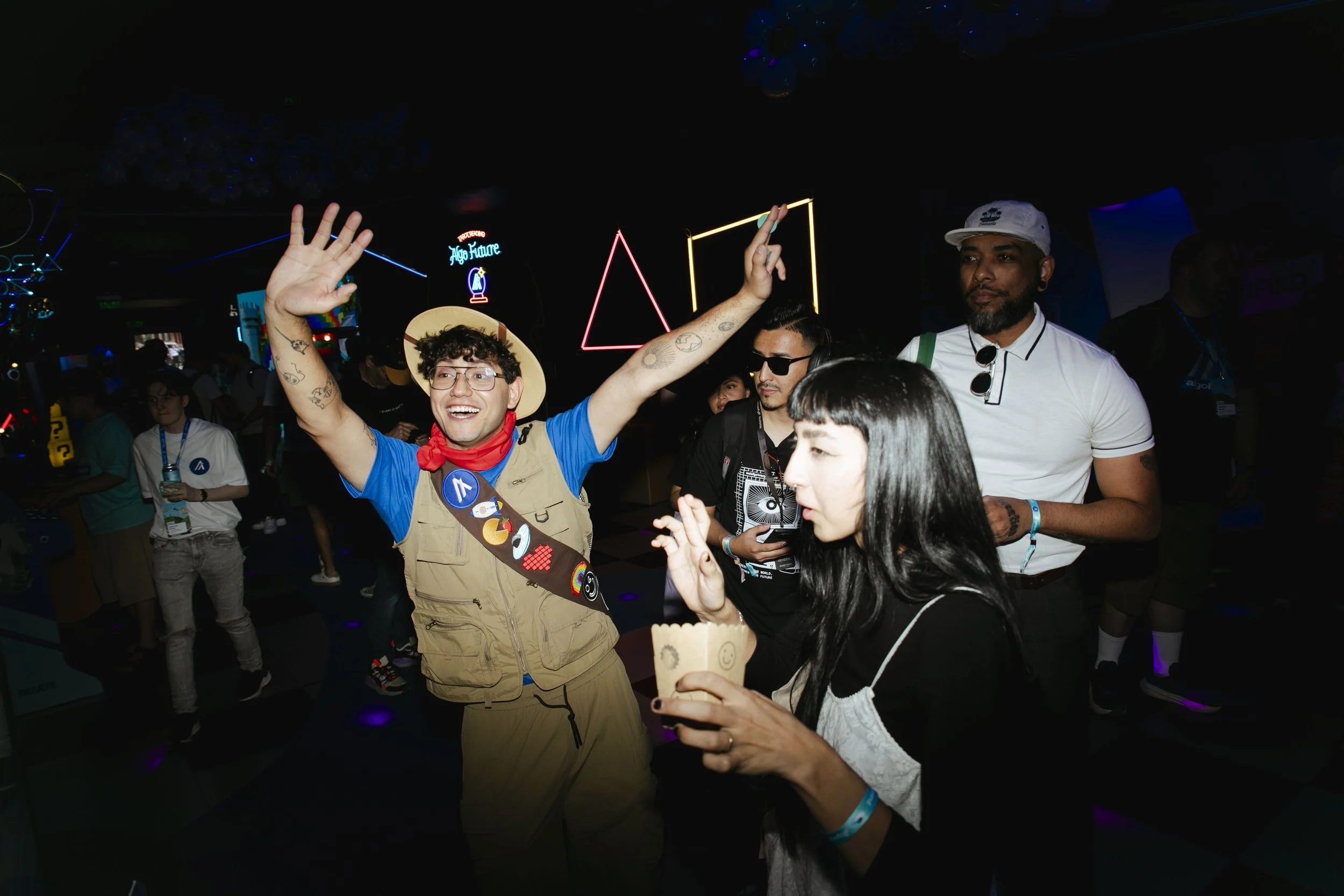 People dancing and socializing at a lively event or party in a dark room with colorful neon lights and geometric decorations.