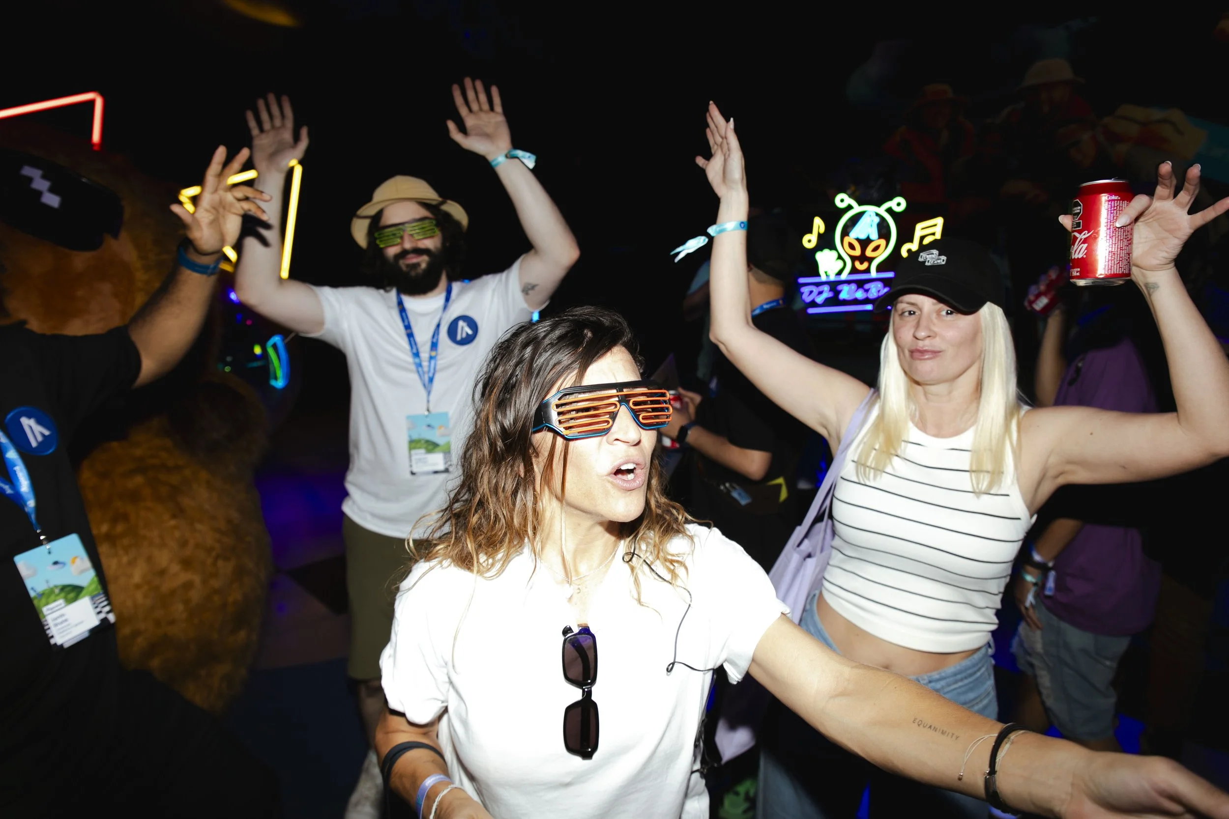 People dancing and celebrating at a party or concert, some wearing neon glasses and hats, one woman holding a soda, colorful neon lights in the background.