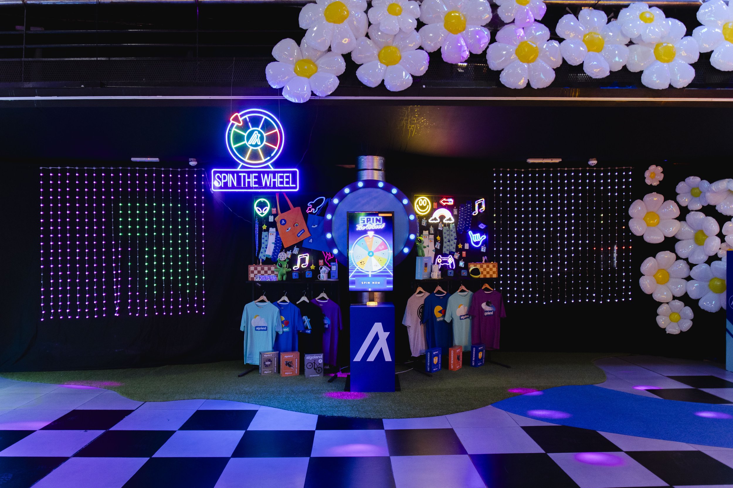 Colorful neon signs and decorations at an indoor arcade or entertainment area, featuring a spinning wheel game, striped shirts hanging on racks, light-up flower decorations on the ceiling, and checkered flooring.