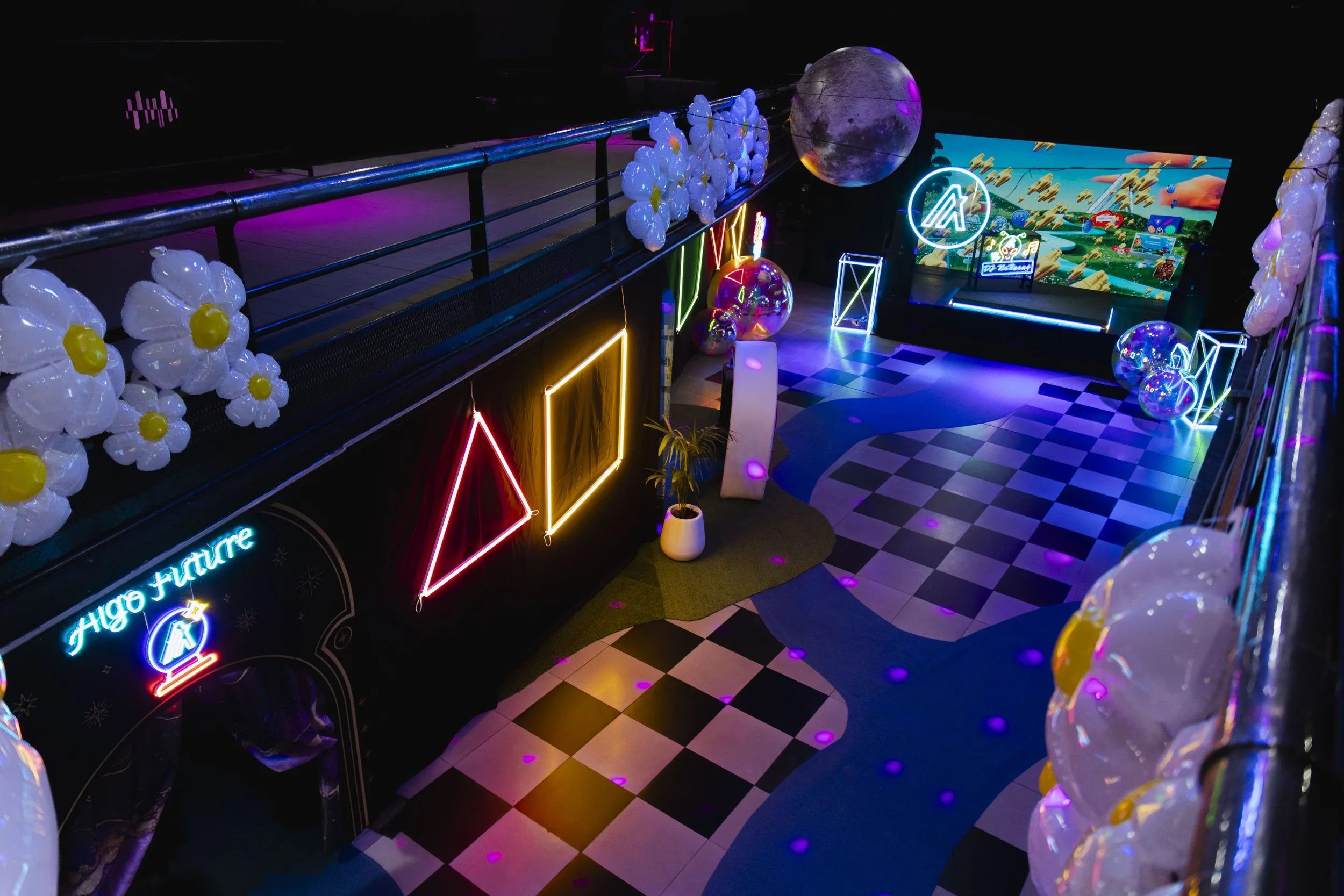 Neon-lit arcade with balloons, geometric shapes, a large moon prop, a digital screen showing a game, and colorful lighting effects.