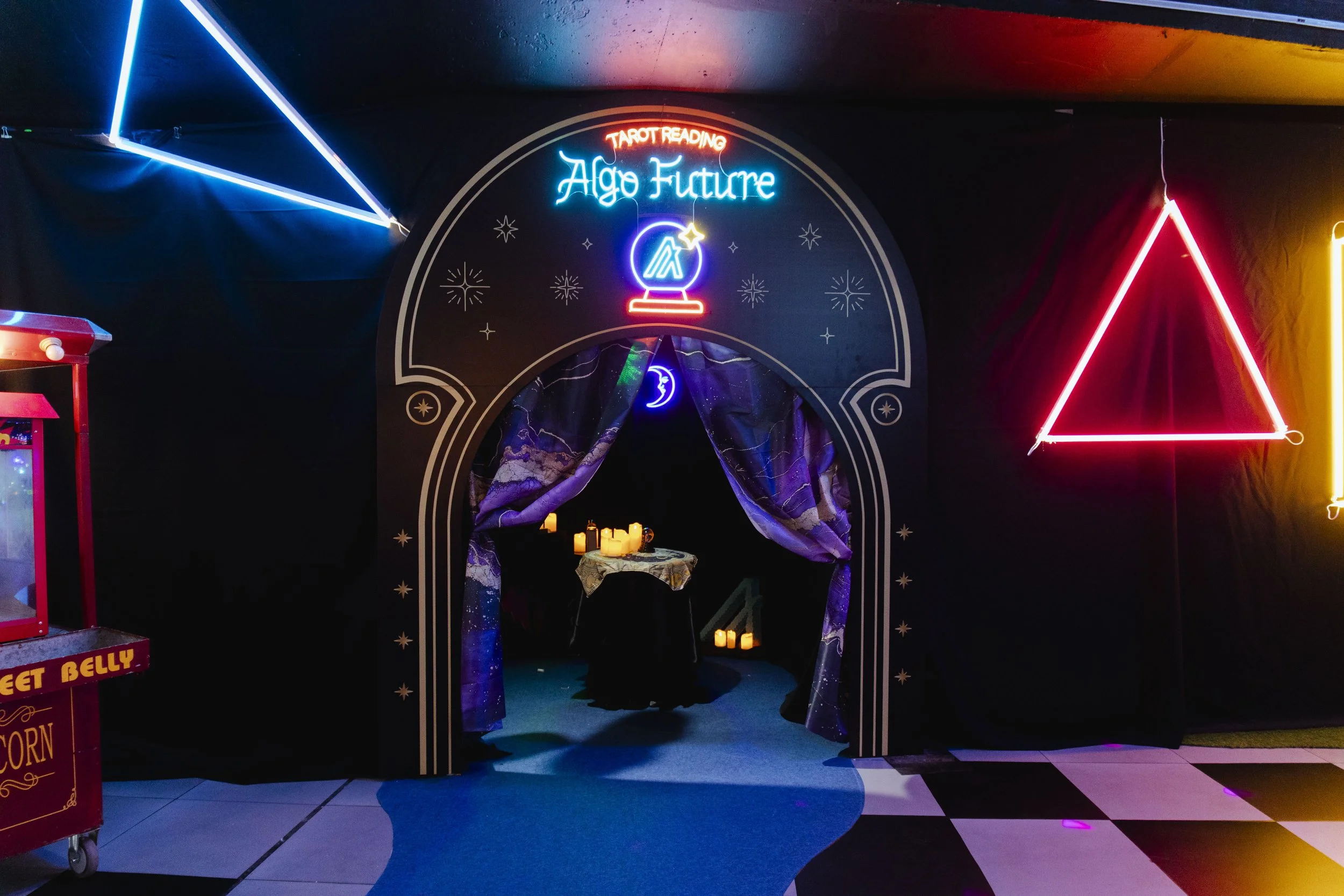 Neon sign reading 'Age Future' above an archway with purple curtains, leading to a table with candles, for a tarot reading setup.