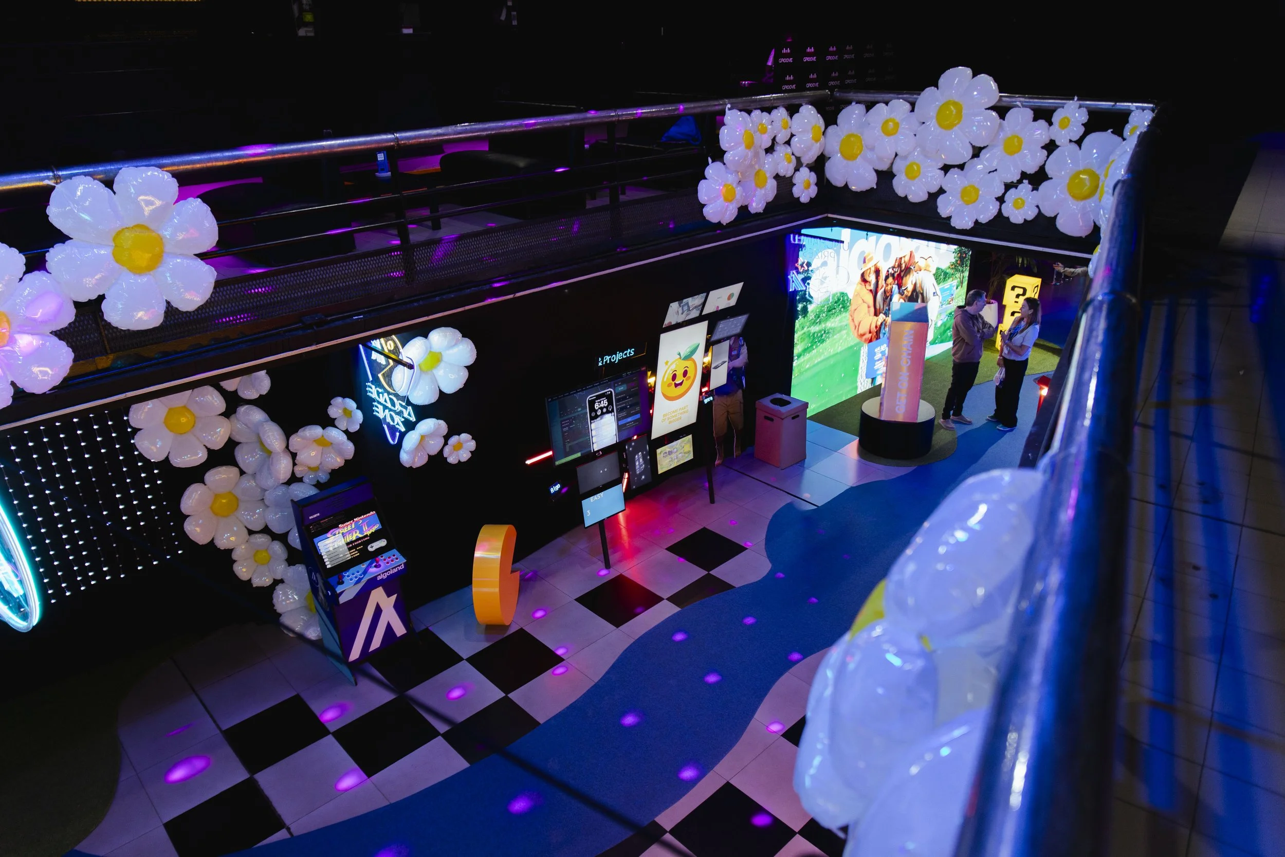 View from an upper level of a colorful indoor space decorated with balloon flowers, digital screens, and interactive displays, with three people standing and talking near a large illuminated graphic and a question mark sign.