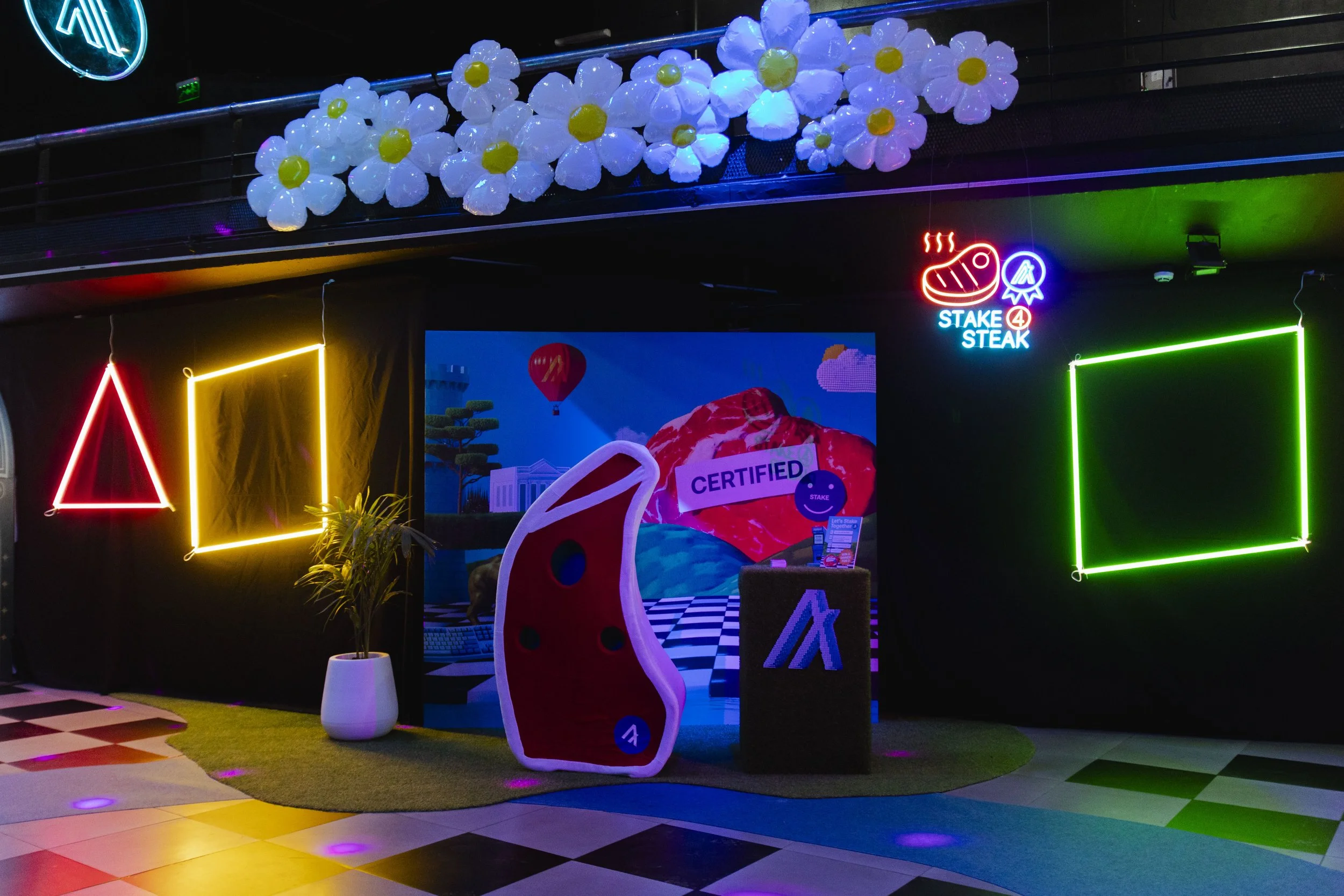 Neon decorated indoor scene with geometric shapes, a potted plant, a digital screen with a scenic background, and a display with a meat cutout labeled 'Certified' at a promotional booth.