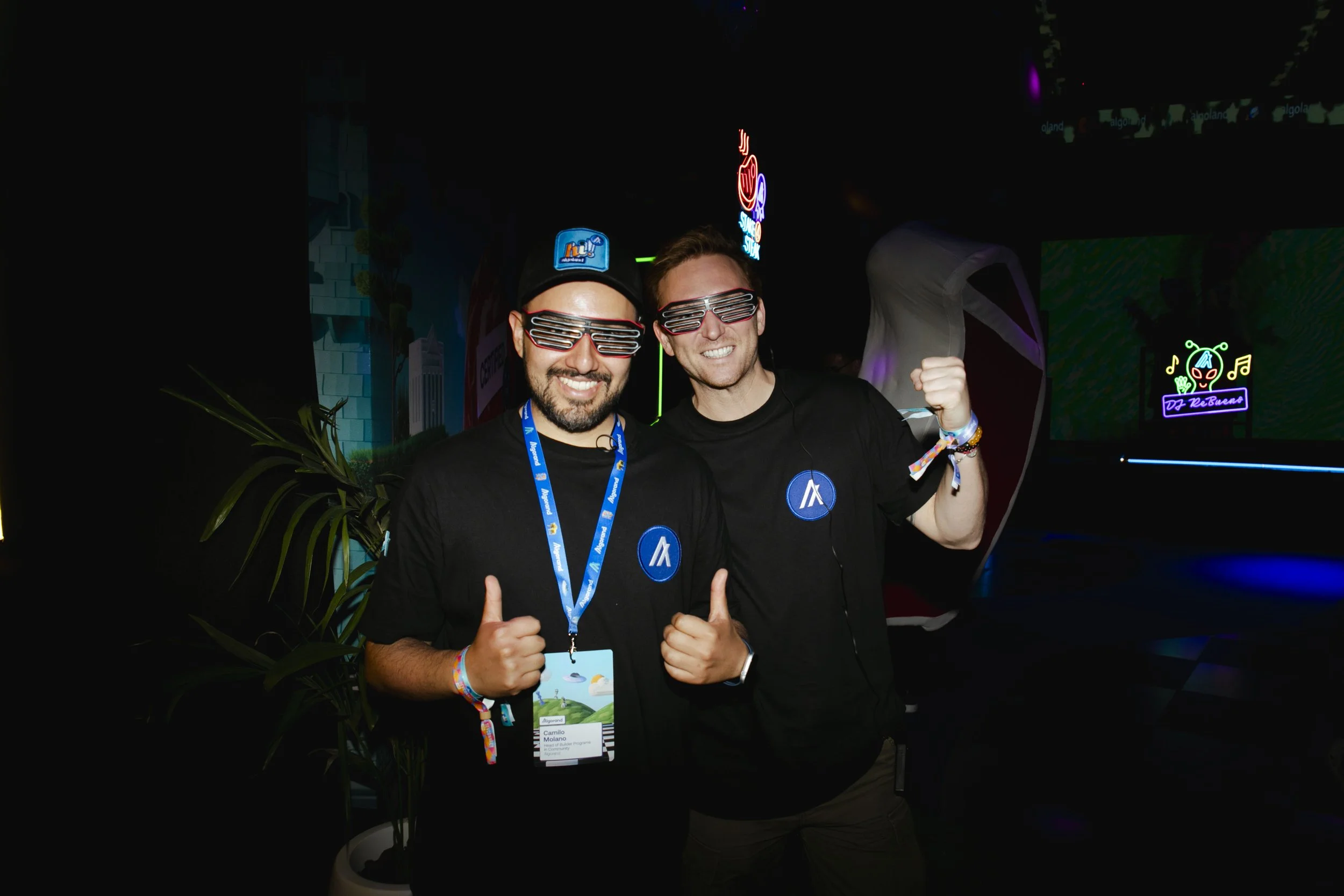 Two men smiling and wearing matching black t-shirts with a blue logo, sunglasses with horizontal stripes, at a neon-lit event, giving thumbs up and showing enthusiasm.