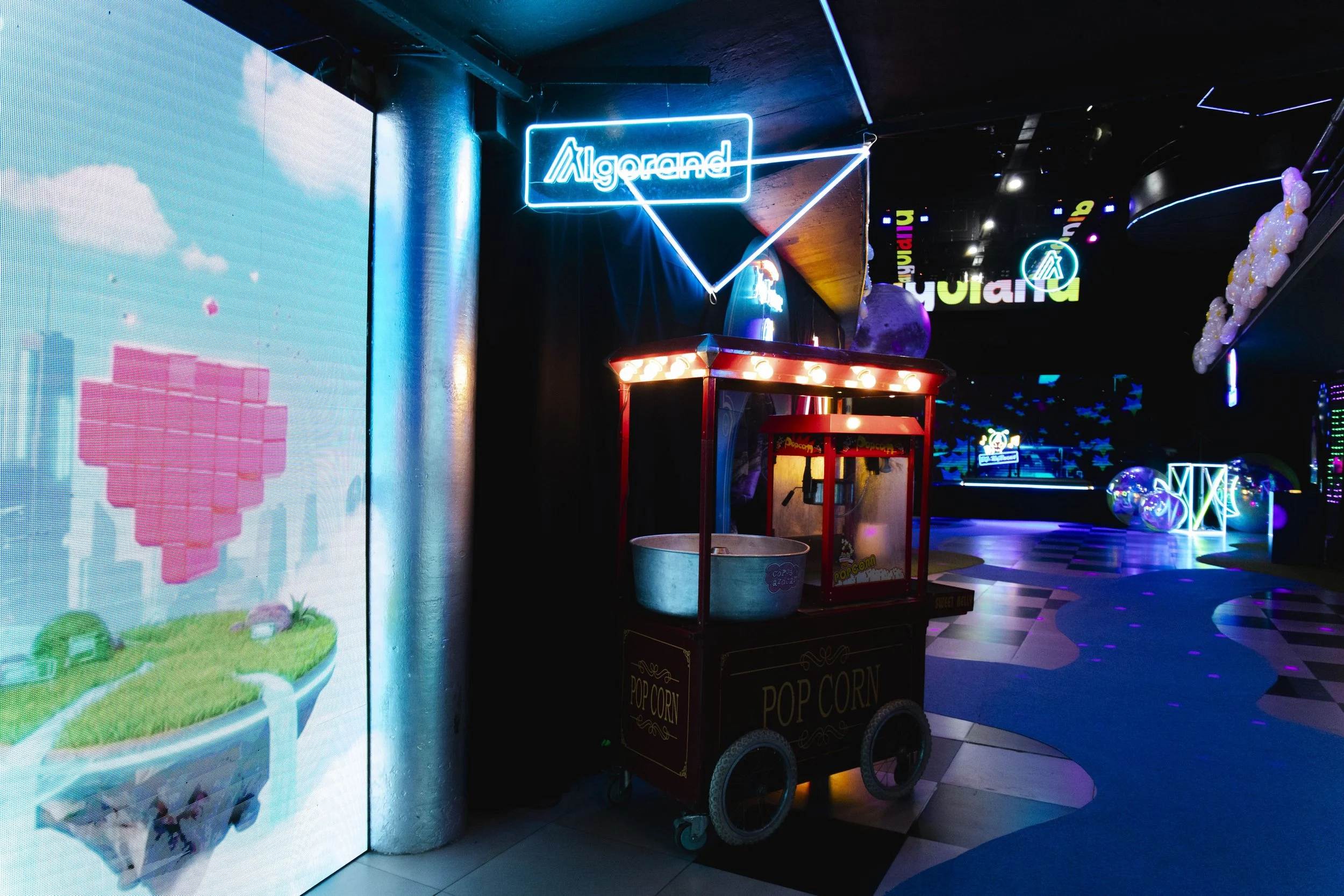 Indoor arcade or entertainment venue with neon signs, purple and blue lighting, and a popcorn stand in the foreground. There is a digital screen on the left showing a pixel art image of a pink heart-shaped character with green surroundings. Neon sign