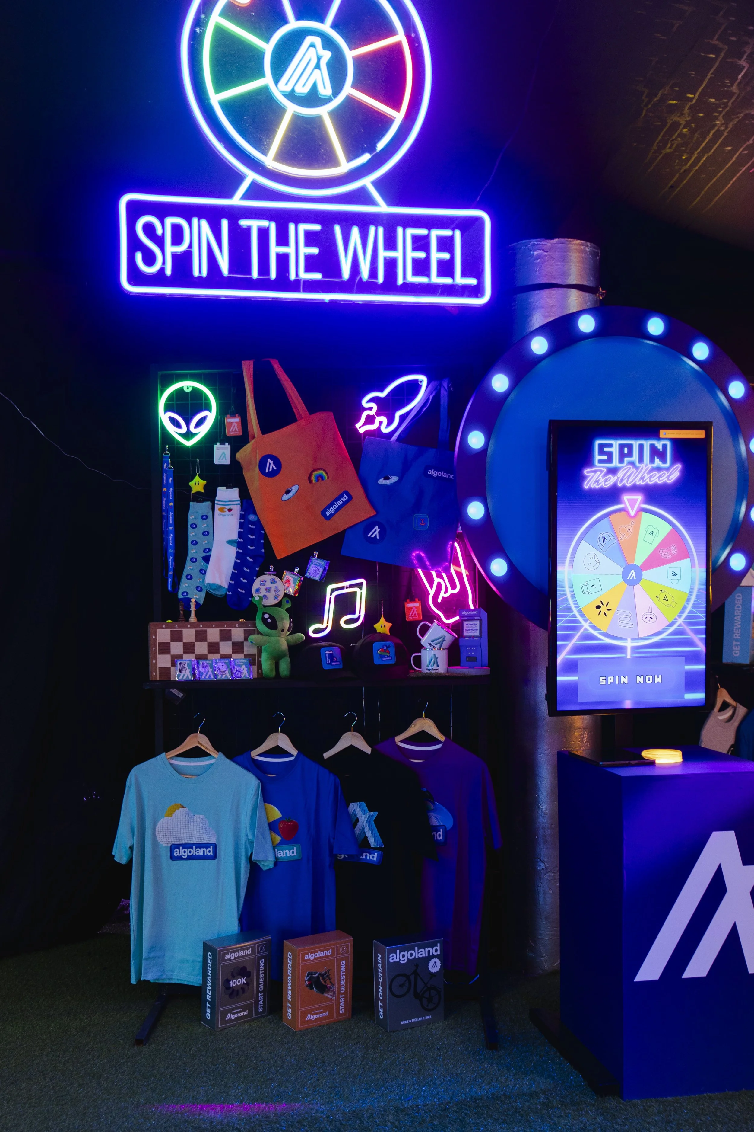 Colorful neon sign that reads "Spin the Wheel," with a prize wheel, merchandise, and t-shirts bearing the "Algoland" logo displayed beneath it.
