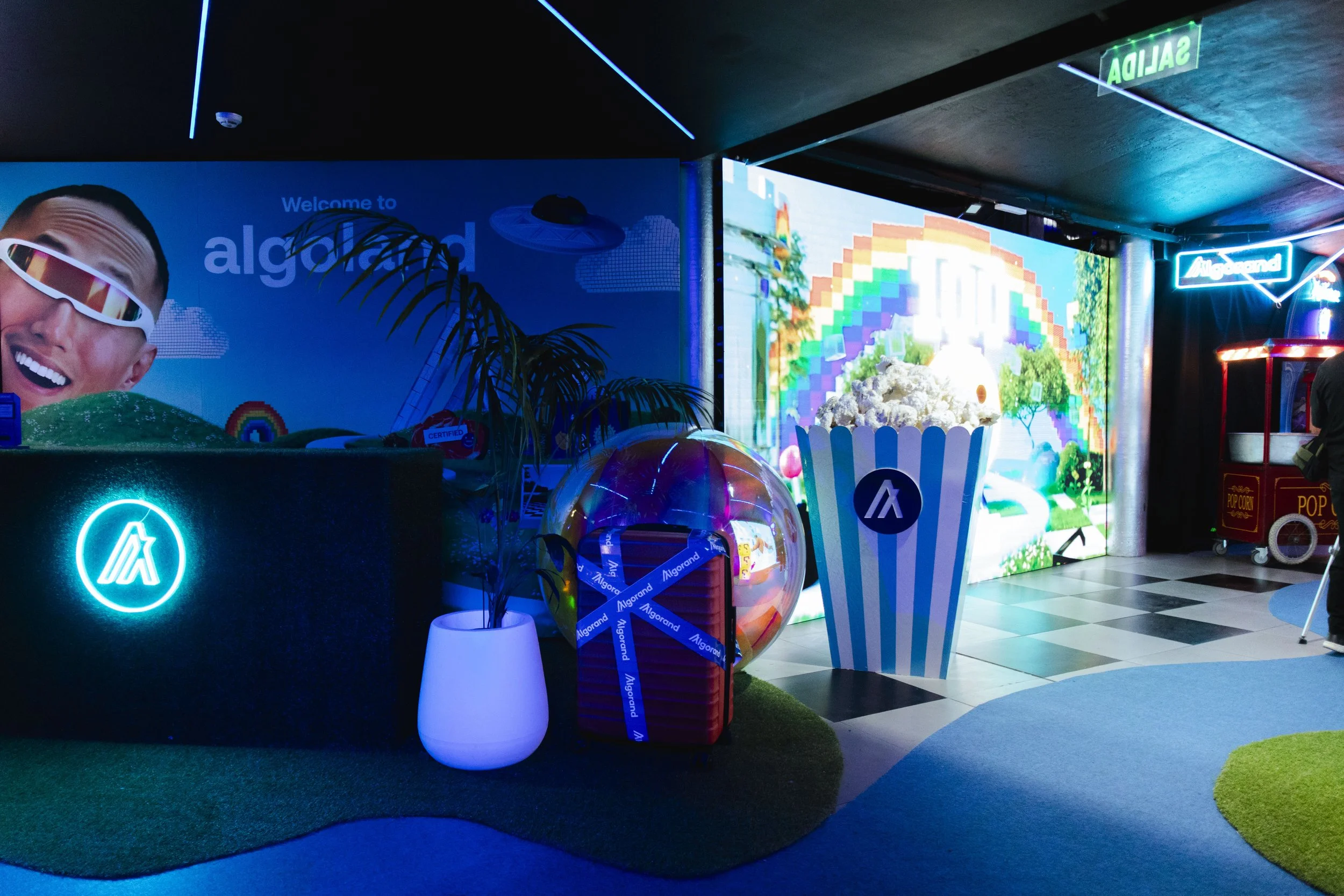 Brightly lit indoor space with neon signs, a large decorative popcorn container, a potted plant, and a colorful digital screen in the background. There is a large branded striped container resembling pop corn container and a small popcorn cart on the