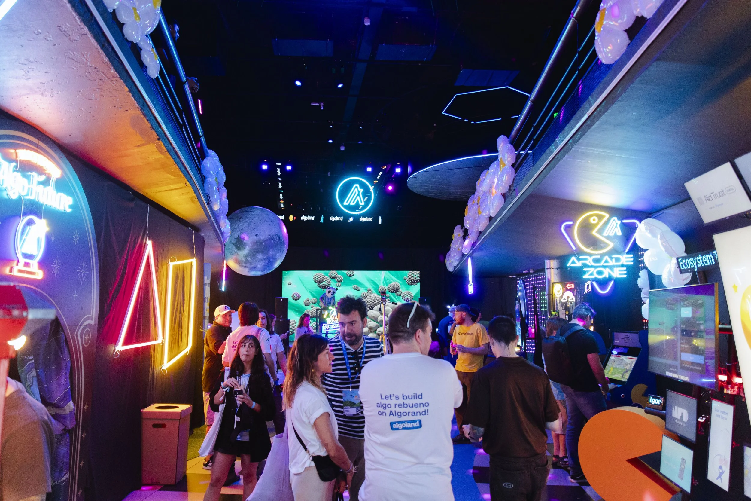 People gathered in a neon-lit indoor arcade or gaming zone decorated with space-themed and digital artwork, including neon signs and hanging white balloons, with screens displaying game interfaces.