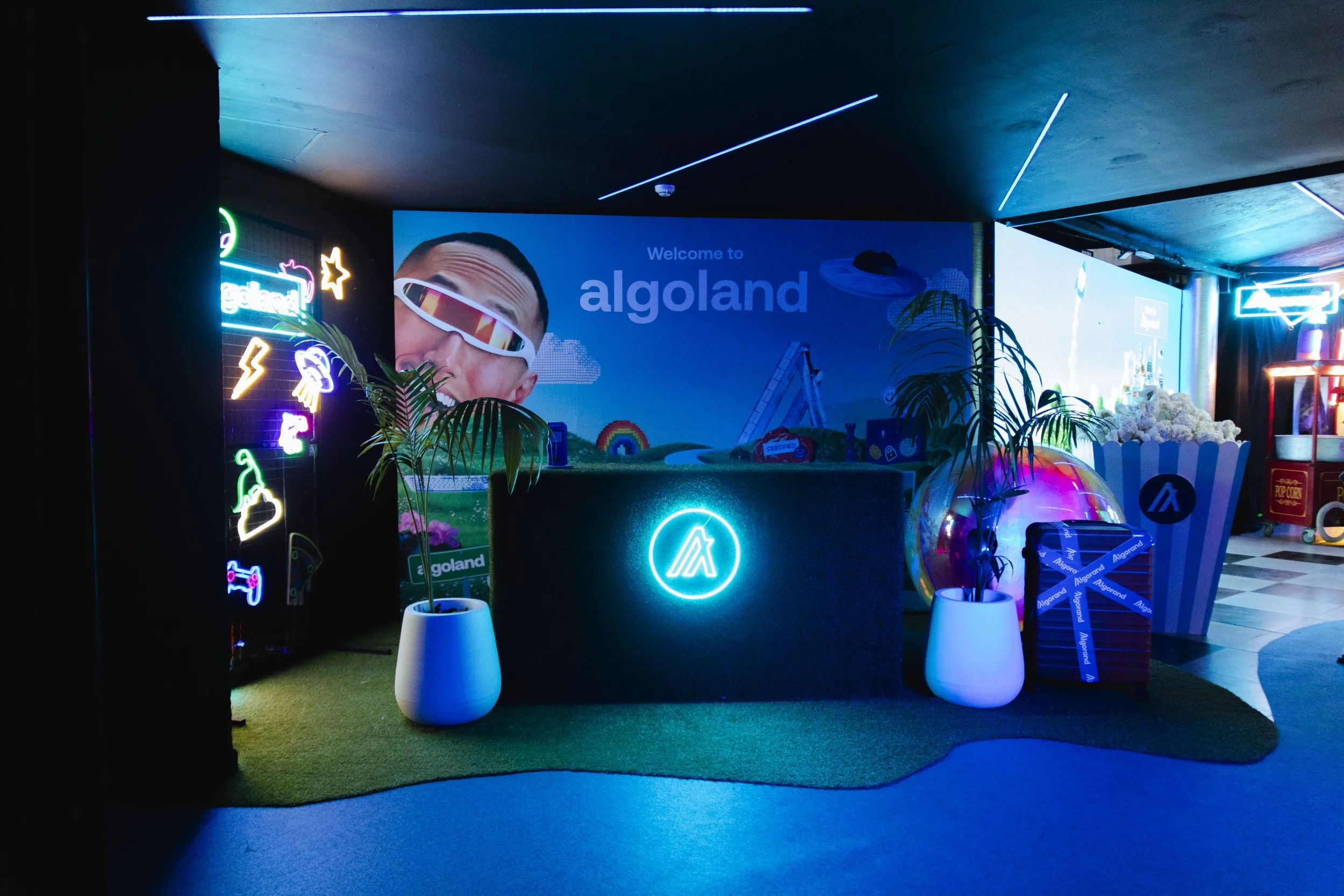 Neon-lit welcome area with a large sign saying 'Welcome to algoland', featuring a person with sunglasses, decorative neon icons, potted plants, and a popcorn container.