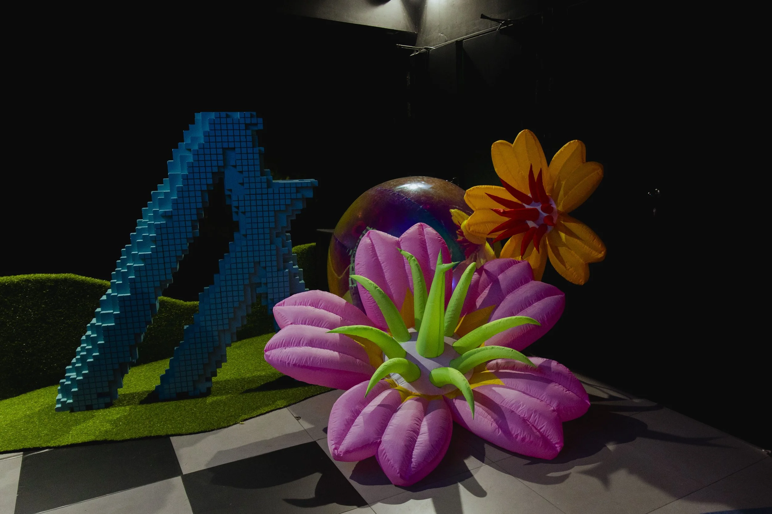 Colorful display with large, pink, and yellow artificial flowers, a reflective sphere, and a blue pixelated dolphin sculpture on a green base.