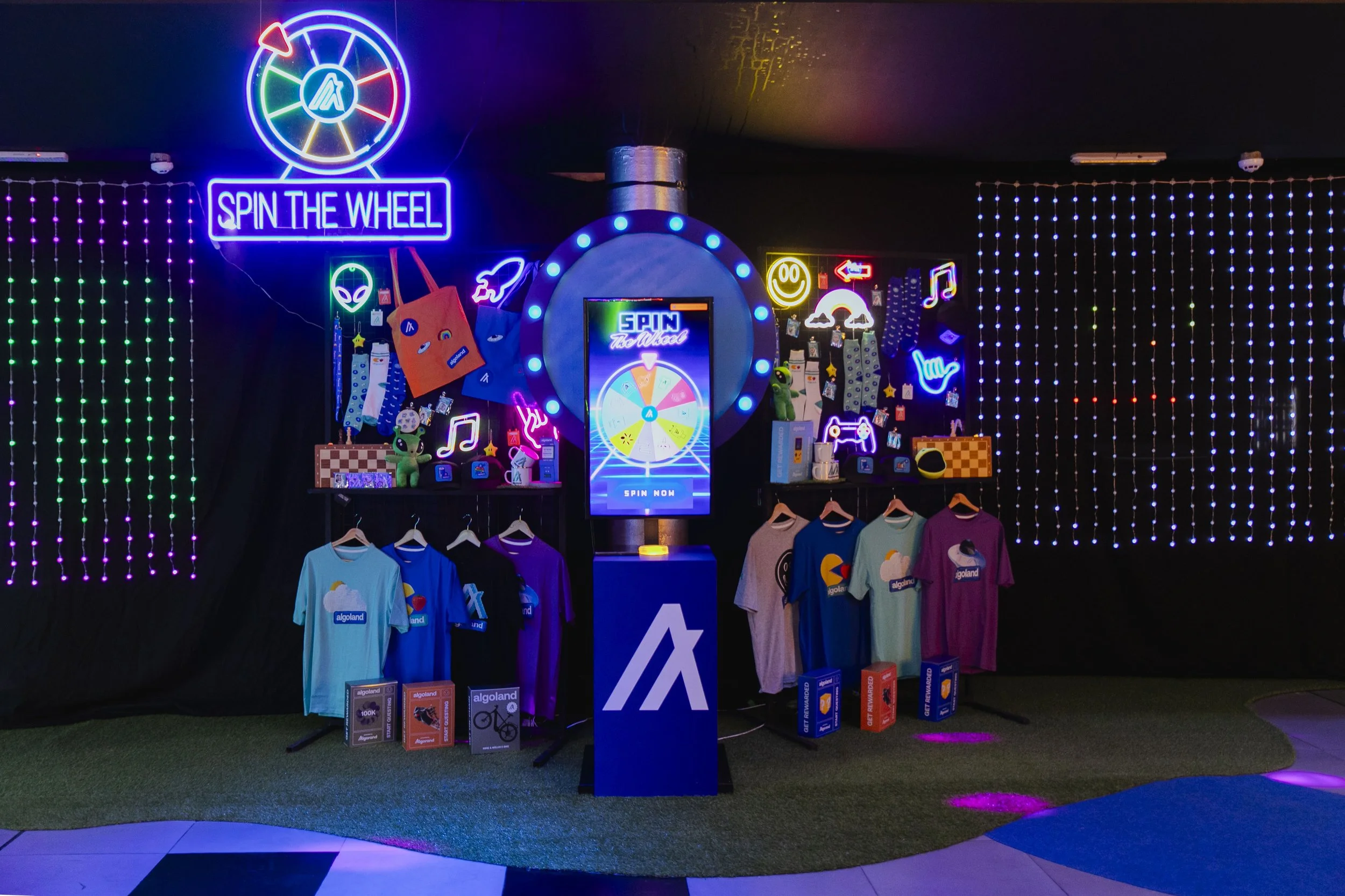 Colorful neon-lit display with a spinning wheel game, clothing items, and various themed merchandise on shelves and hangers in a dark room decorated with LED lights and glowing signs.