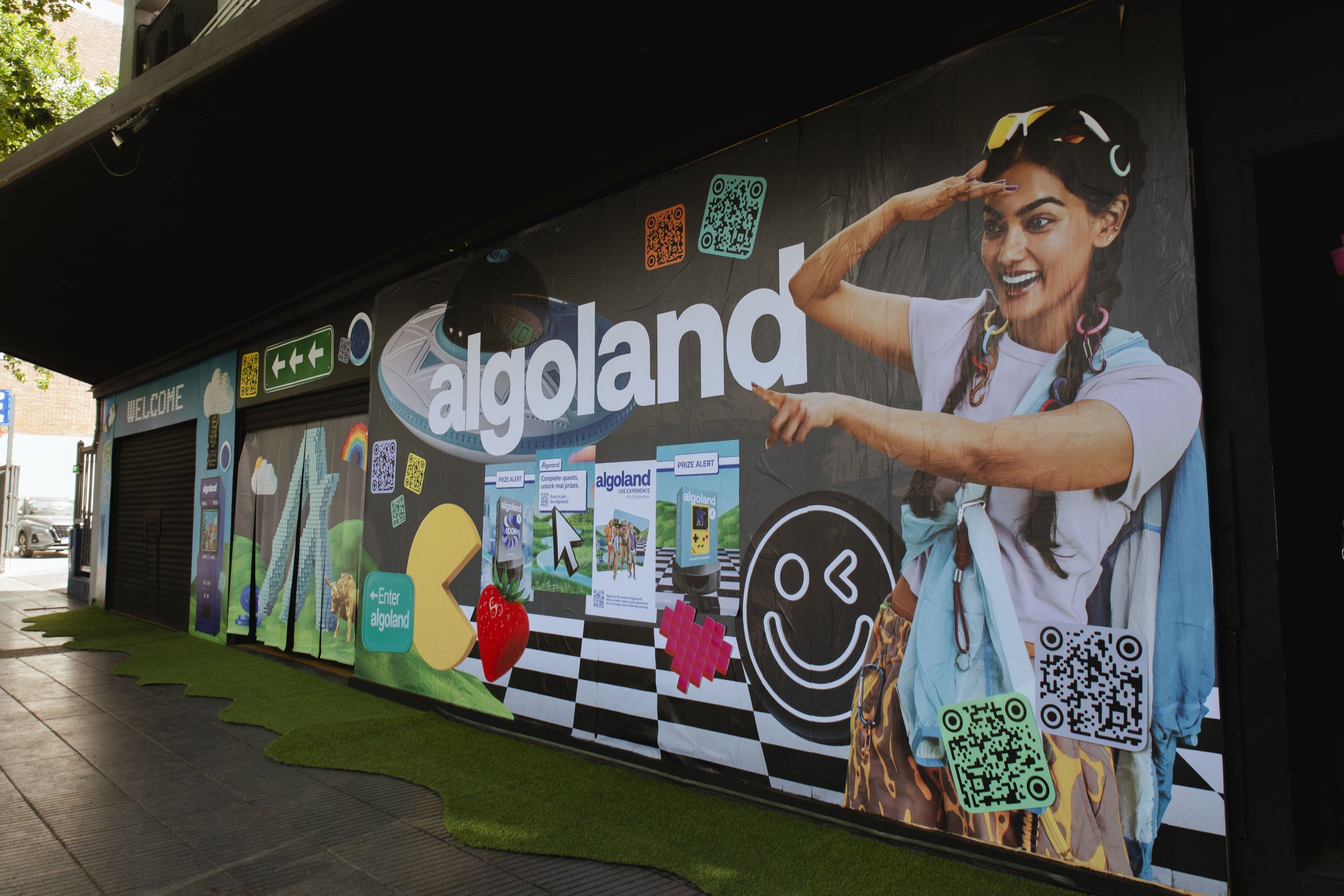 Colorful advertisement for a theme park called Algoland featuring a woman with dark hair wearing sunglasses and colorful accessories, smiling and pointing, with various playful graphics, QR codes, and signage.