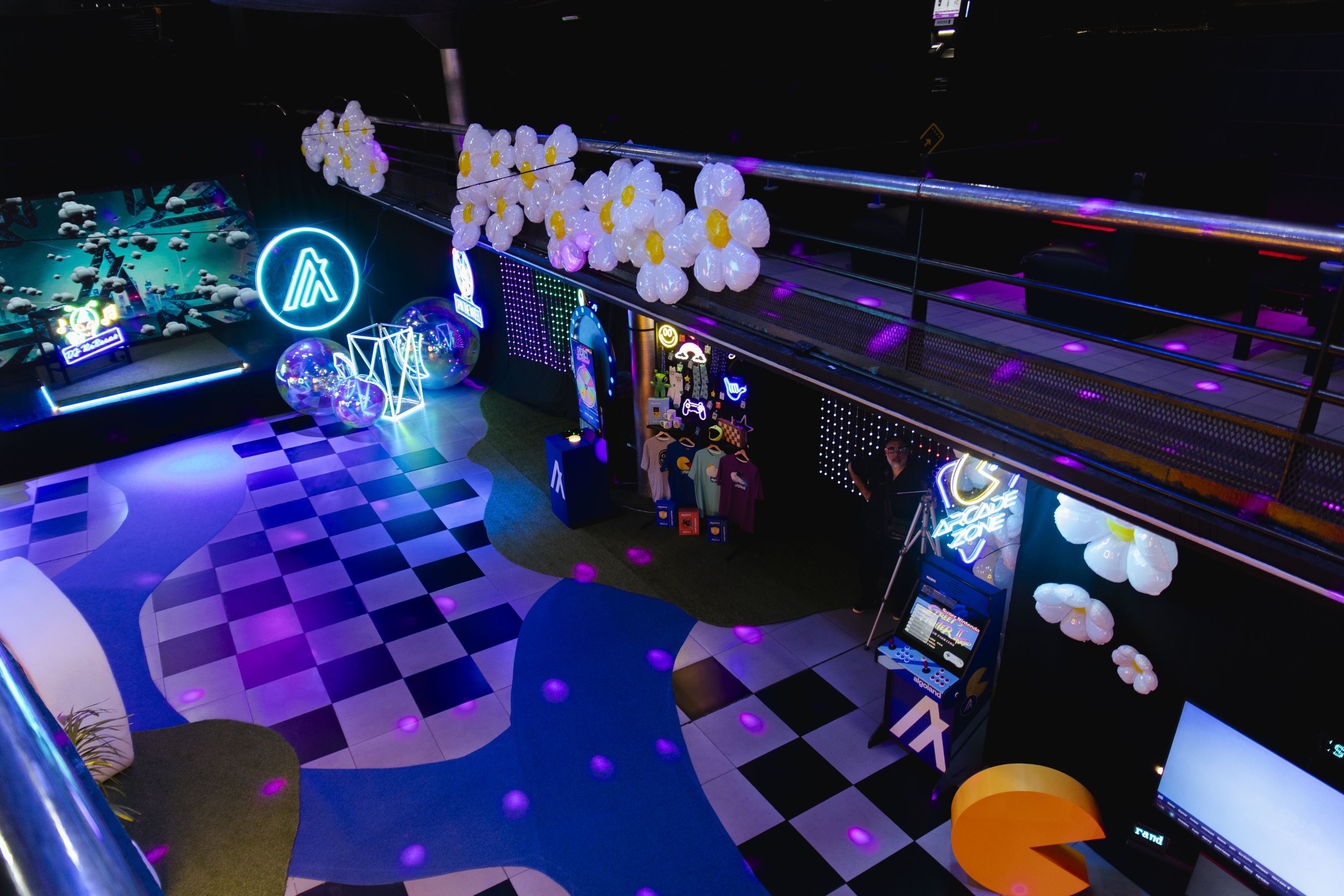 A colorful indoor arcade or gaming zone with neon lights, gaming paraphernalia, and a patterned floor with purple and pink lighting.