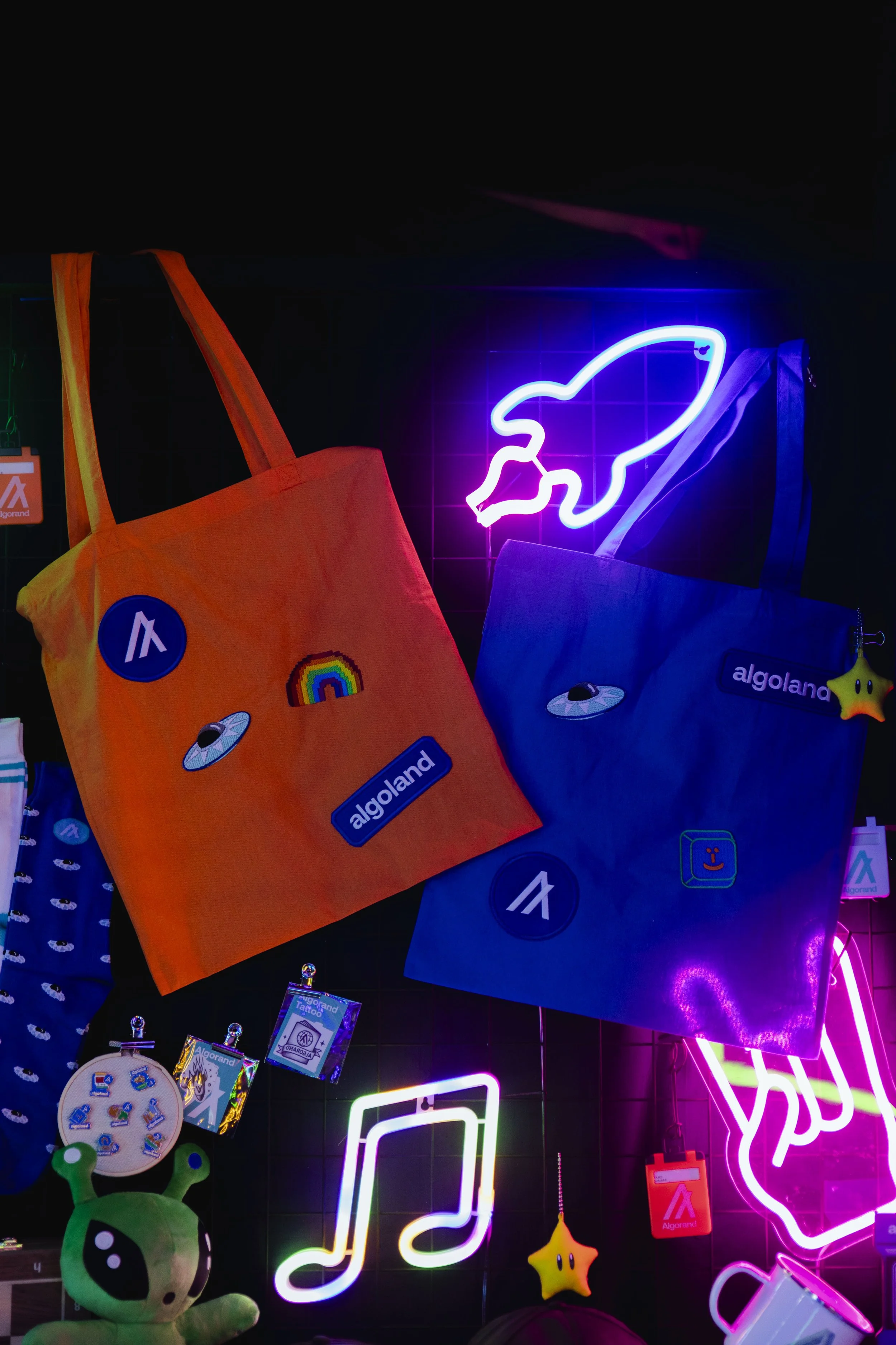 Colorful tote bags with stickers and patches hanging against a black wall with neon signs, including a rocket, musical notes, and stars.