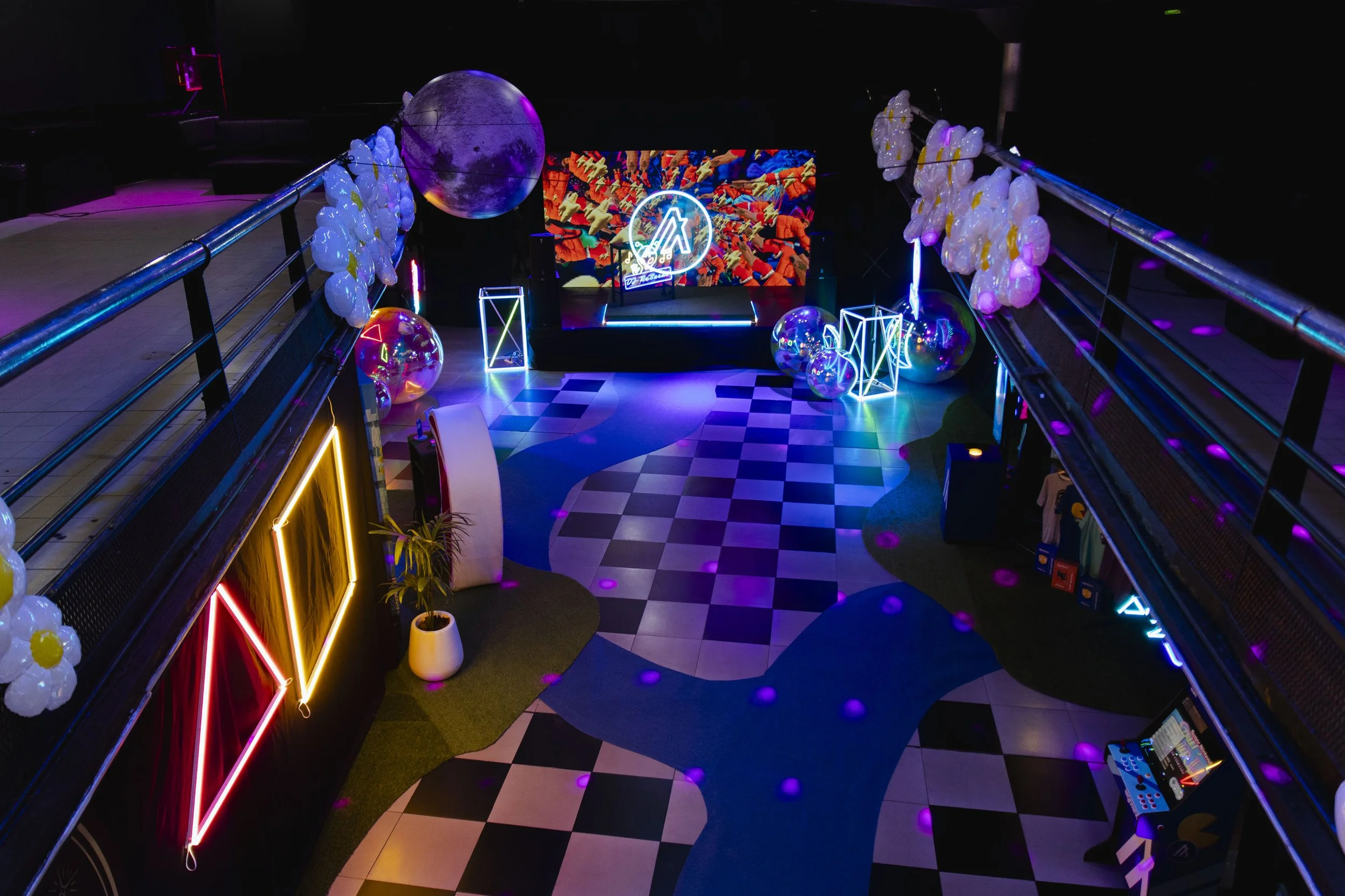 Colorful interior with neon lights, large balloons, a digital screen displaying vibrant abstract art, a dance floor with checkered and wavy patterns, and a stage with a circular neon sign.