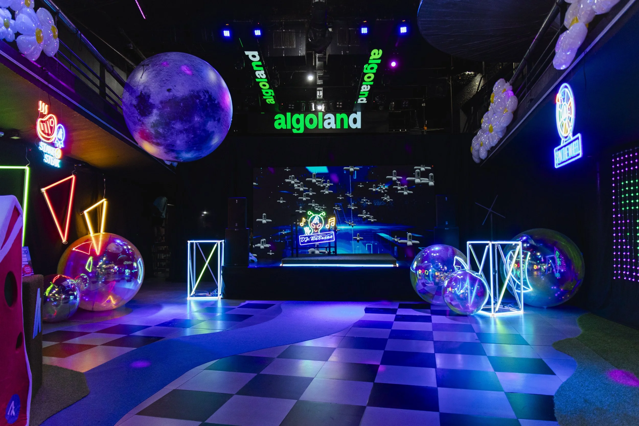 Neon-lit indoor space at Algoland, featuring large holographic globes, neon geometric shapes, a checkered floor, LED signs, and a stage with a digital screen background and musical icons.