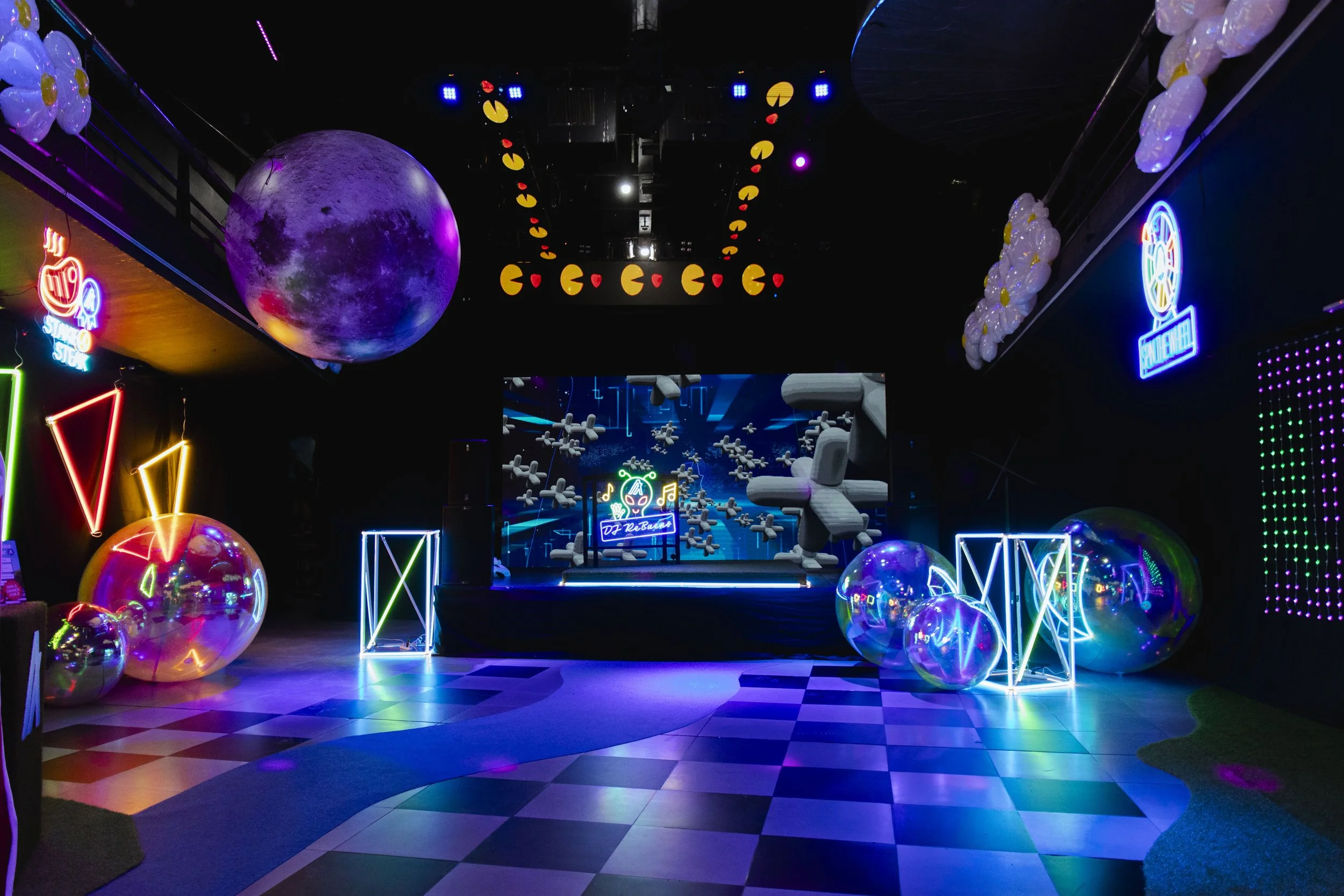 An indoor entertainment venue decorated with neon signs, large reflective balloons, and Pac-Man-themed decor. The room has a checkered floor and a stage with a digital display, among colorful, glowing geometric shapes and moon decorations.