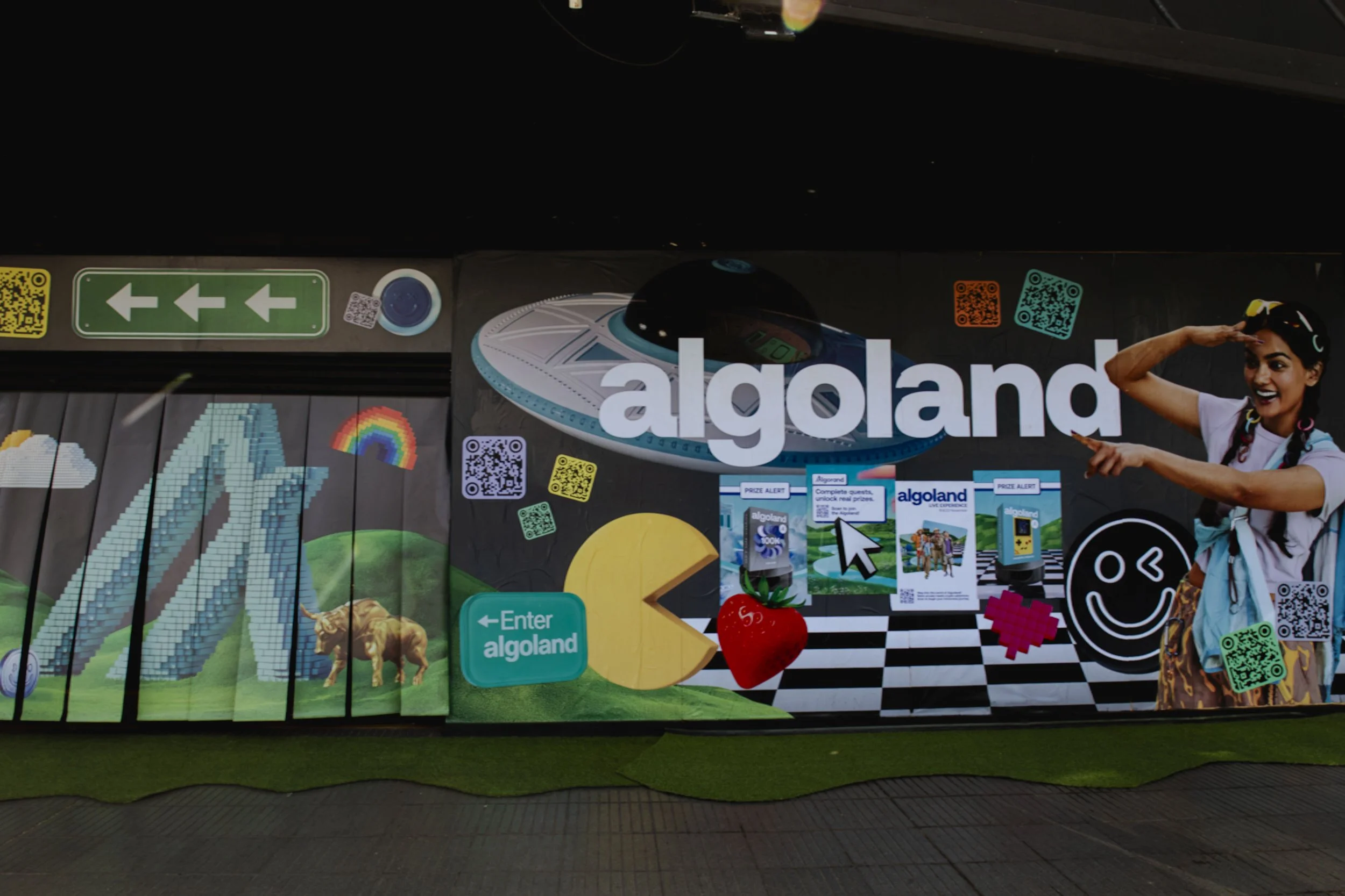 Colorful advertising mural for Algoland, featuring a spaceship, a pixelated mountain, a rainbow, a strawberry, and a cheerful young woman pointing and saluting, with QR codes and various playful graphics.