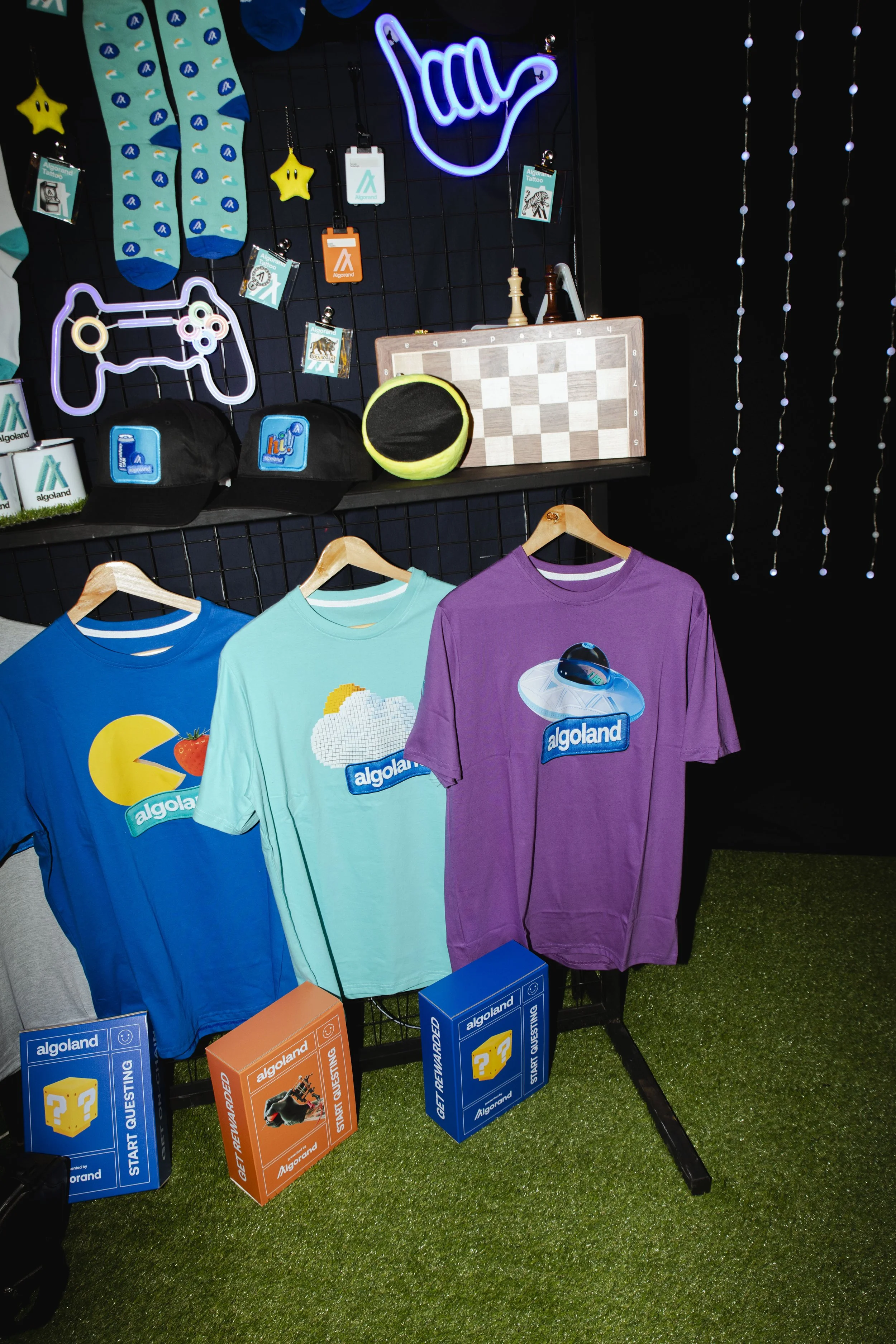 Display of colorful T-shirts with brand logos, two black caps, neon signs, game controller image, chessboard, plush ball, and various merchandise and decorations on a black grid wall and green artificial grass floor.