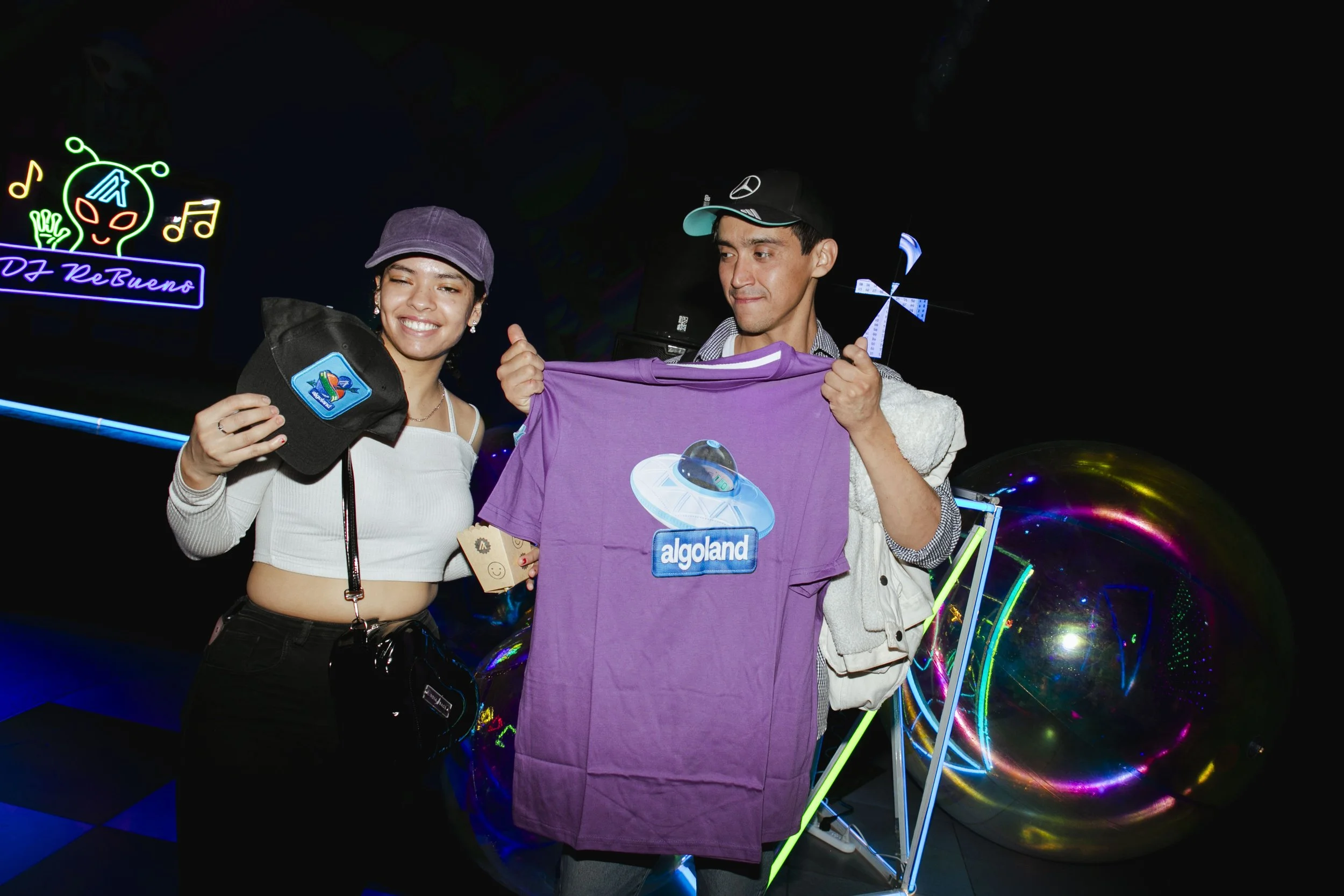 A young woman and man at an indoor arcade or entertainment venue. The woman is smiling and holding a black cap with a colorful patch, while the man is holding up a purple T-shirt with an 'Algolando' spaceship logo. Neon signs and colorful light displ