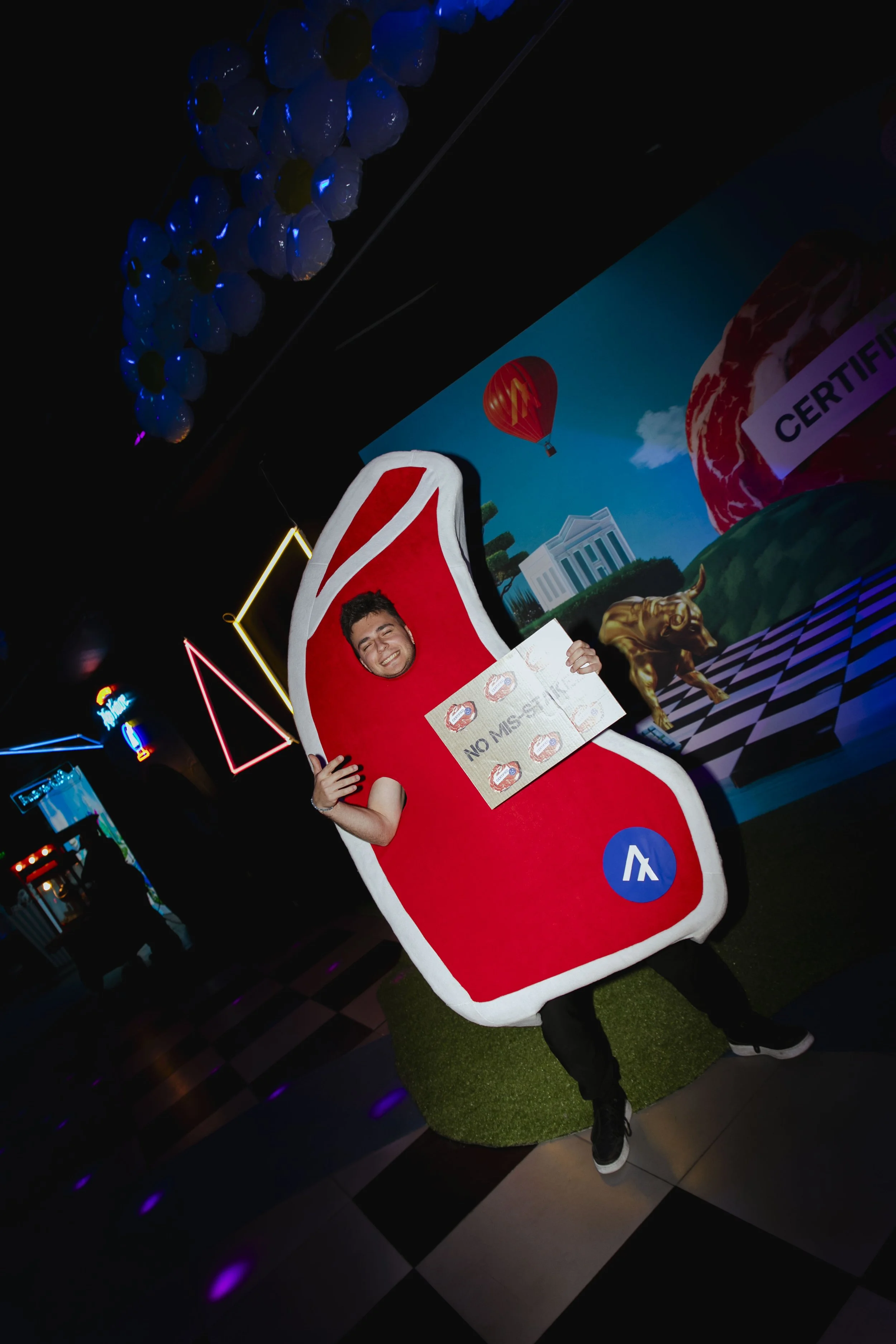 Person in a sushi-shaped costume holding a sign that reads "No Messy" at an indoor event with neon lights and colorful decorations.