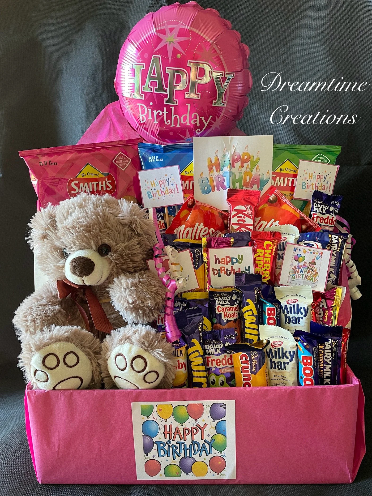 A large pink gift box decorated with a colorful 'Happy Birthday' card, filled with assorted snacks, a plush teddy bear, and birthday decorations.
