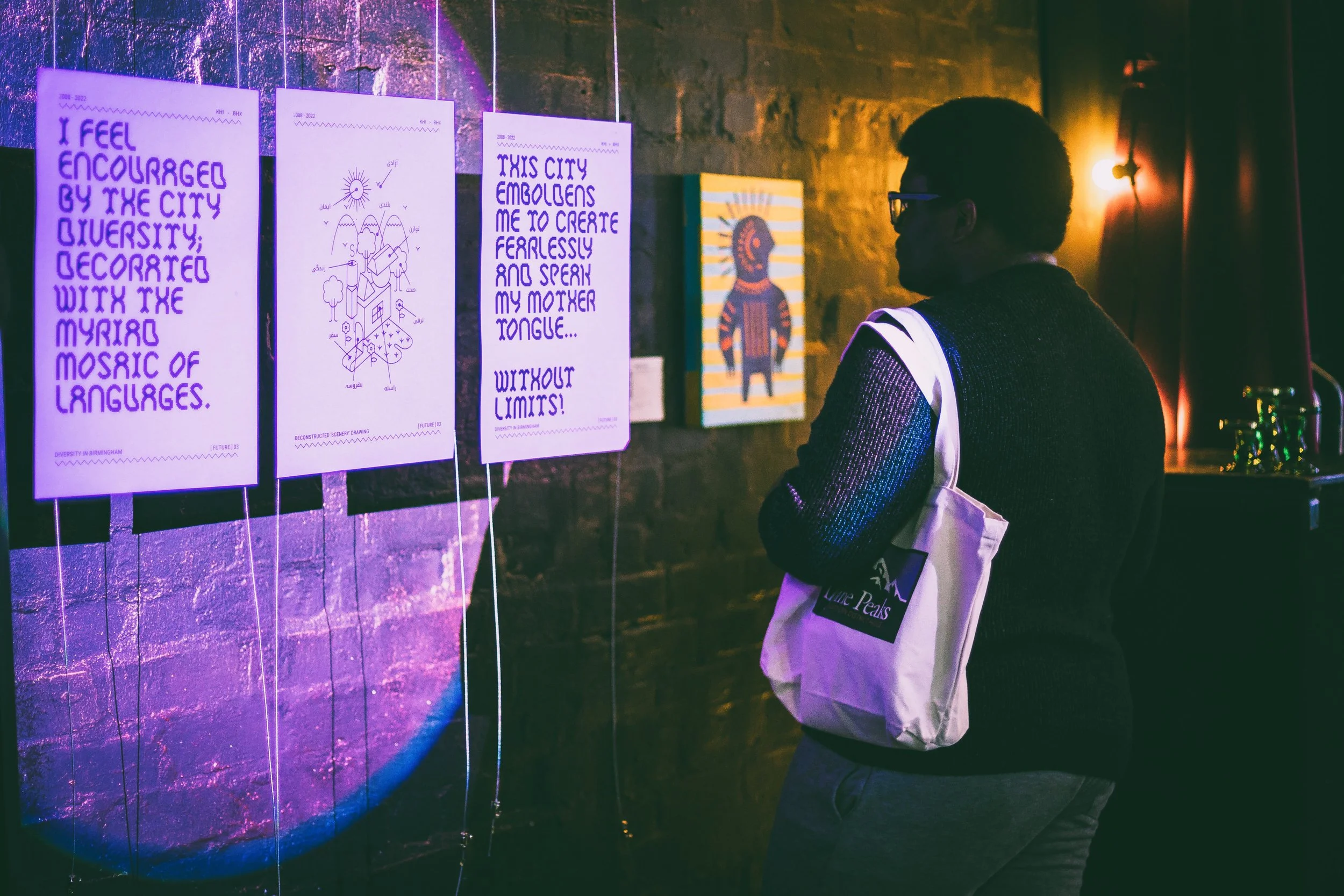 A person with glasses and a white bag looks at art posters displayed on a brick wall in a gallery. The posters include text and drawings, illuminated by colorful lighting.