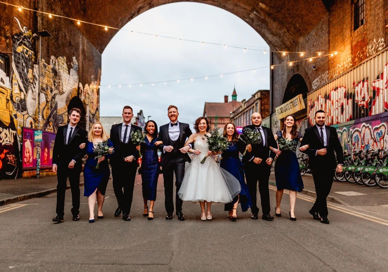 A wedding party of ten people walking arm in arm under a graffiti-covered bridge, dressed in formal attire with the bride in a white gown and bridesmaids wearing blue dresses, celebrating and smiling.