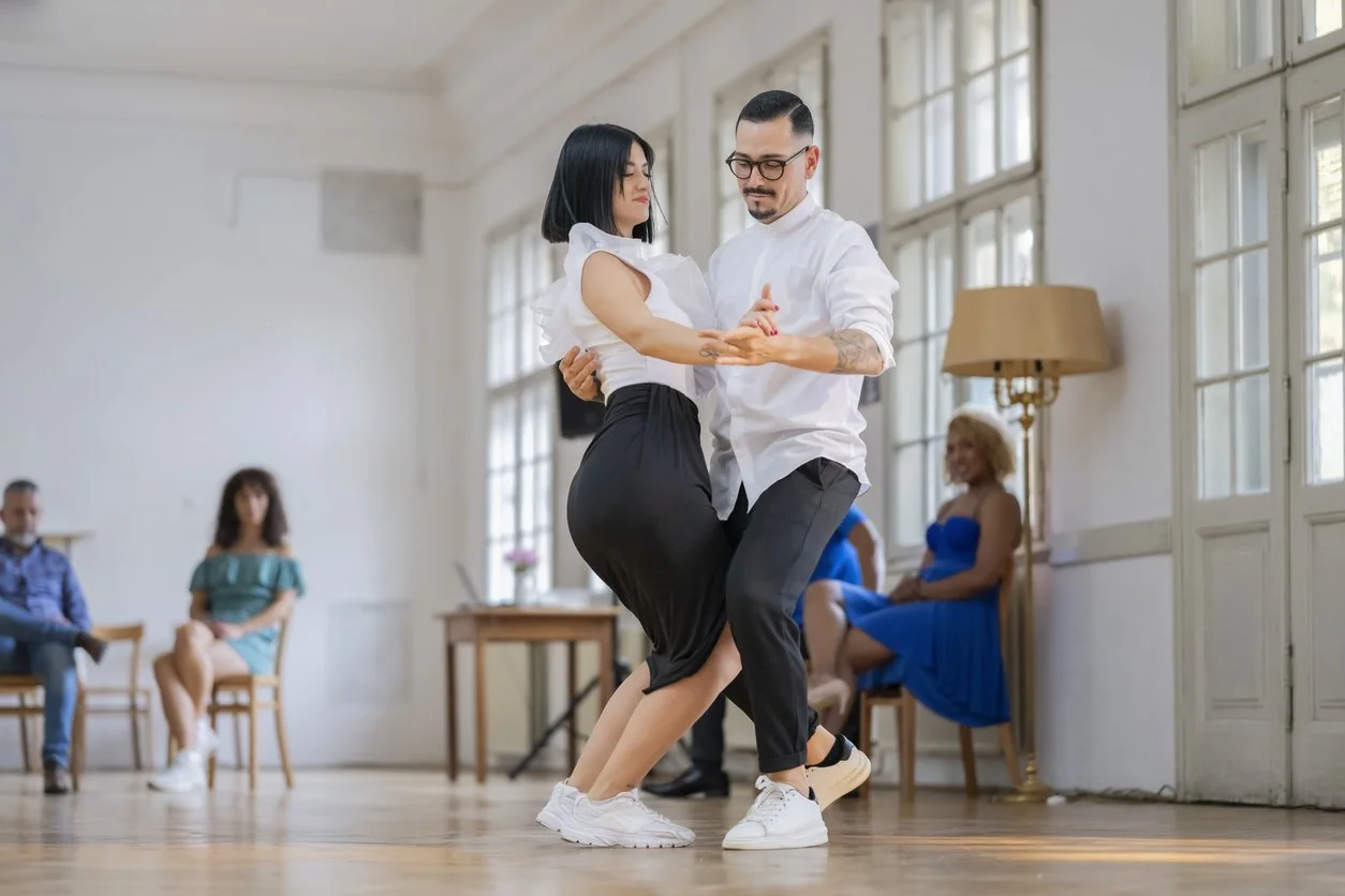 Un couple danse dans une salle lumineuse avec plusieurs spectateurs assis, fenêtres grandes et lampadaire