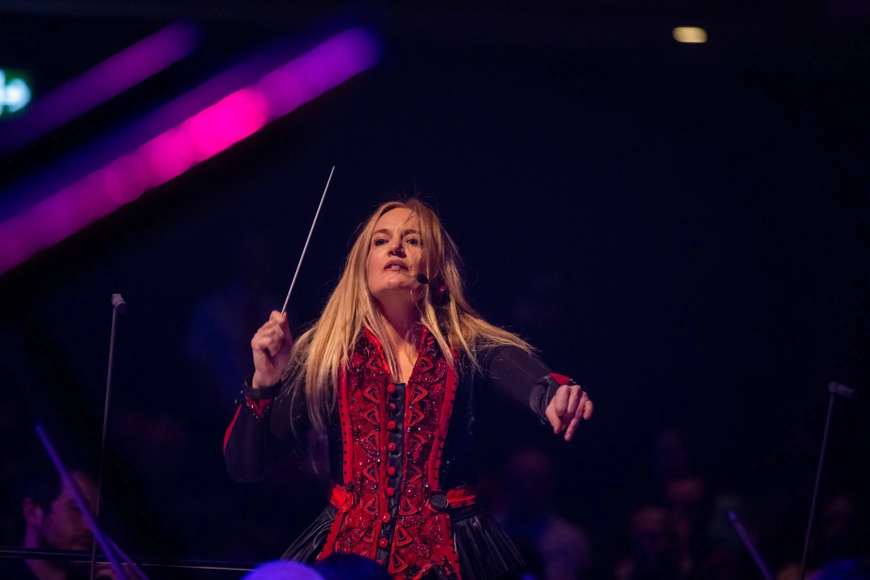 A woman conducting an orchestra during a performance, holding a baton in her right hand, with a dark background and purple and pink stage lights overhead.