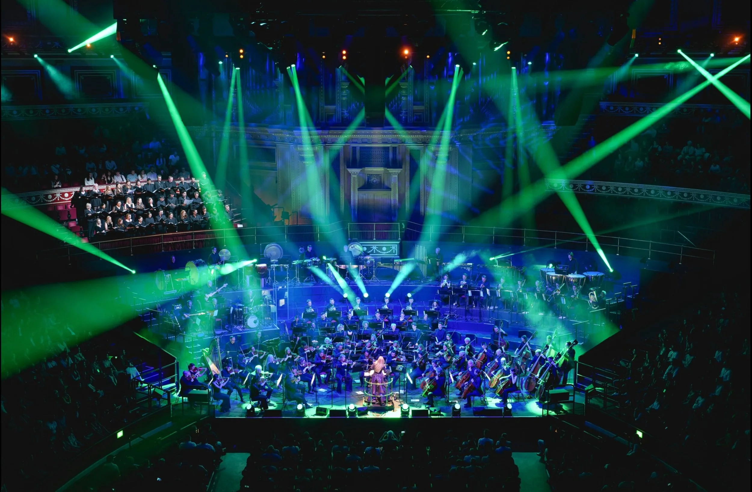 Orchestra performing on stage with green and blue stage lighting, audience seated in a concert hall.