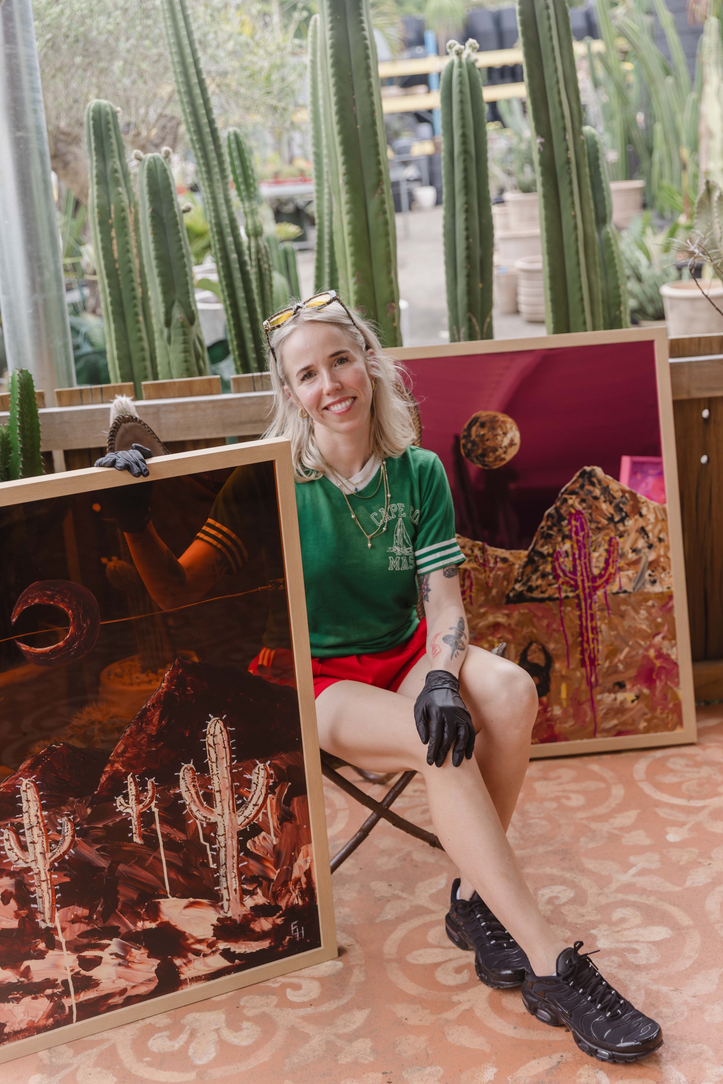 Emily Handlin, contemporary Australian artist, seated with framed desert and cactus artworks inspired by surreal landscapes and the Australian outback.