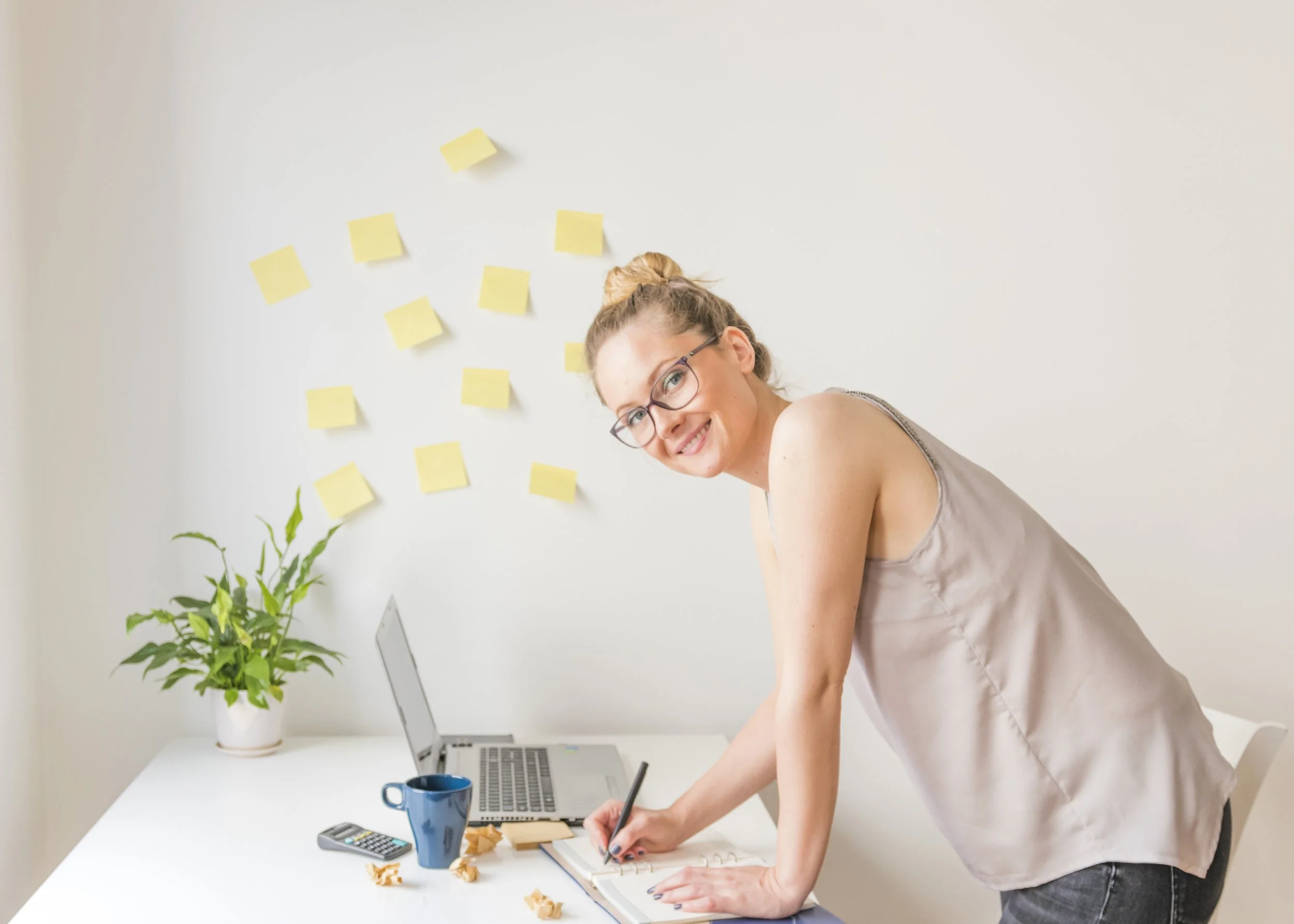 Woman leaning over a desk, writing in a notebook with a laptop, blue mug, and calculator. Yellow sticky notes on the wall and a potted plant are visible.