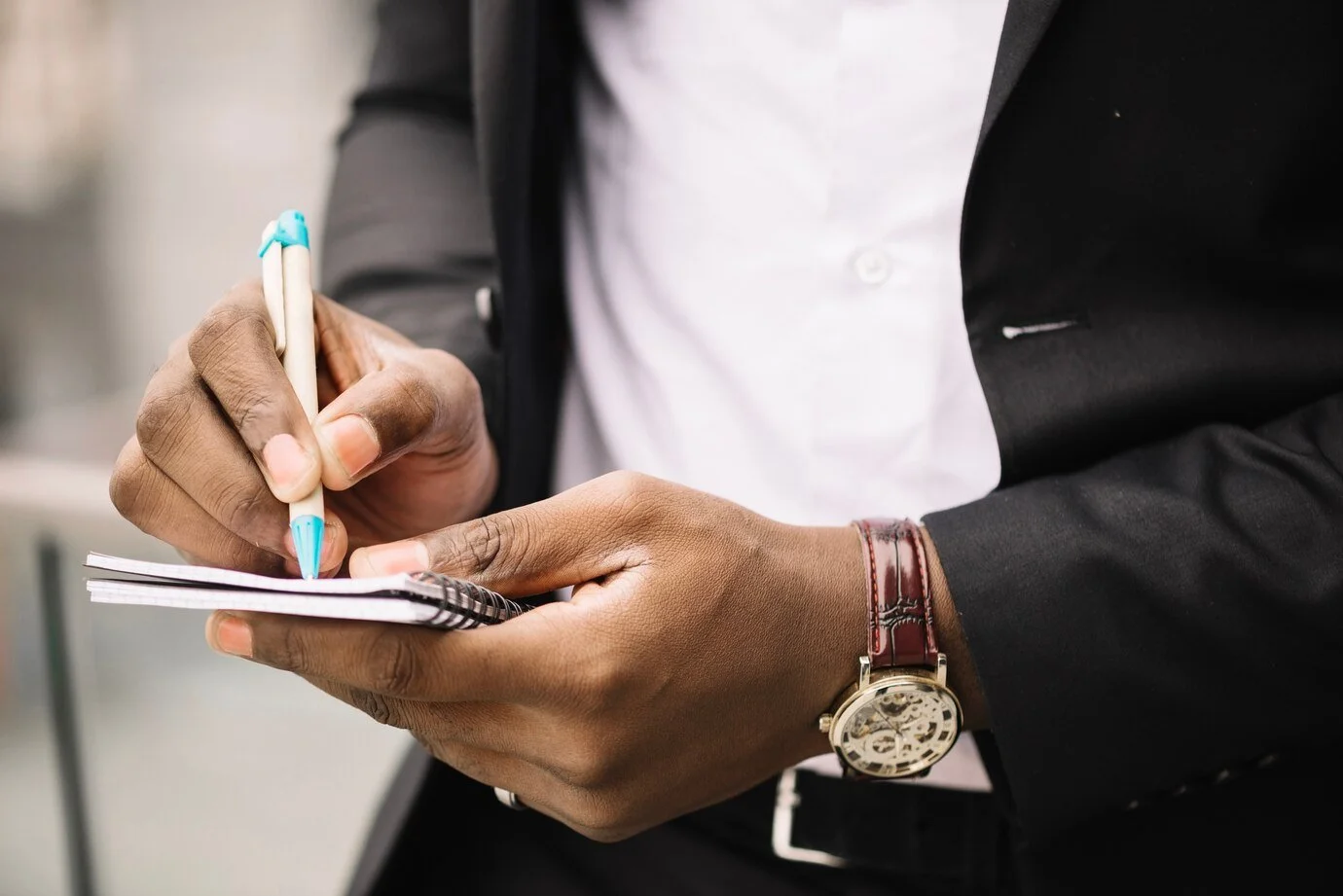 Close-up of a person in a suit writing in a notebook with a pen, wearing a wristwatch.