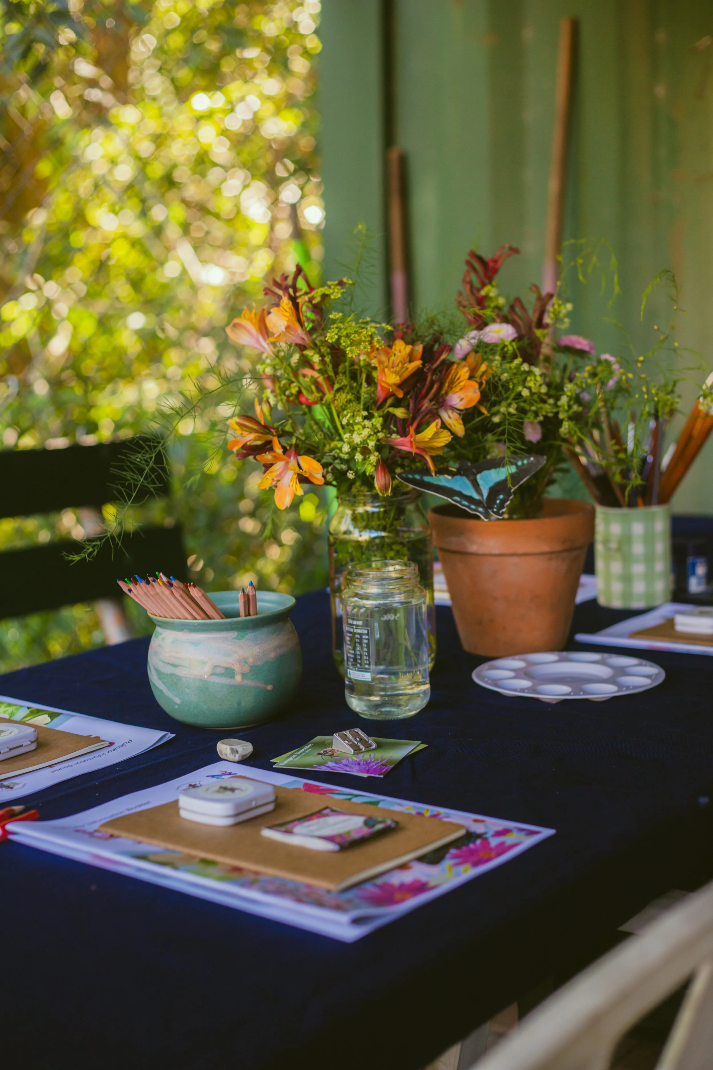 A table with a floral centerpiece, potted plants, and art supplies, set outdoors with a green background.
