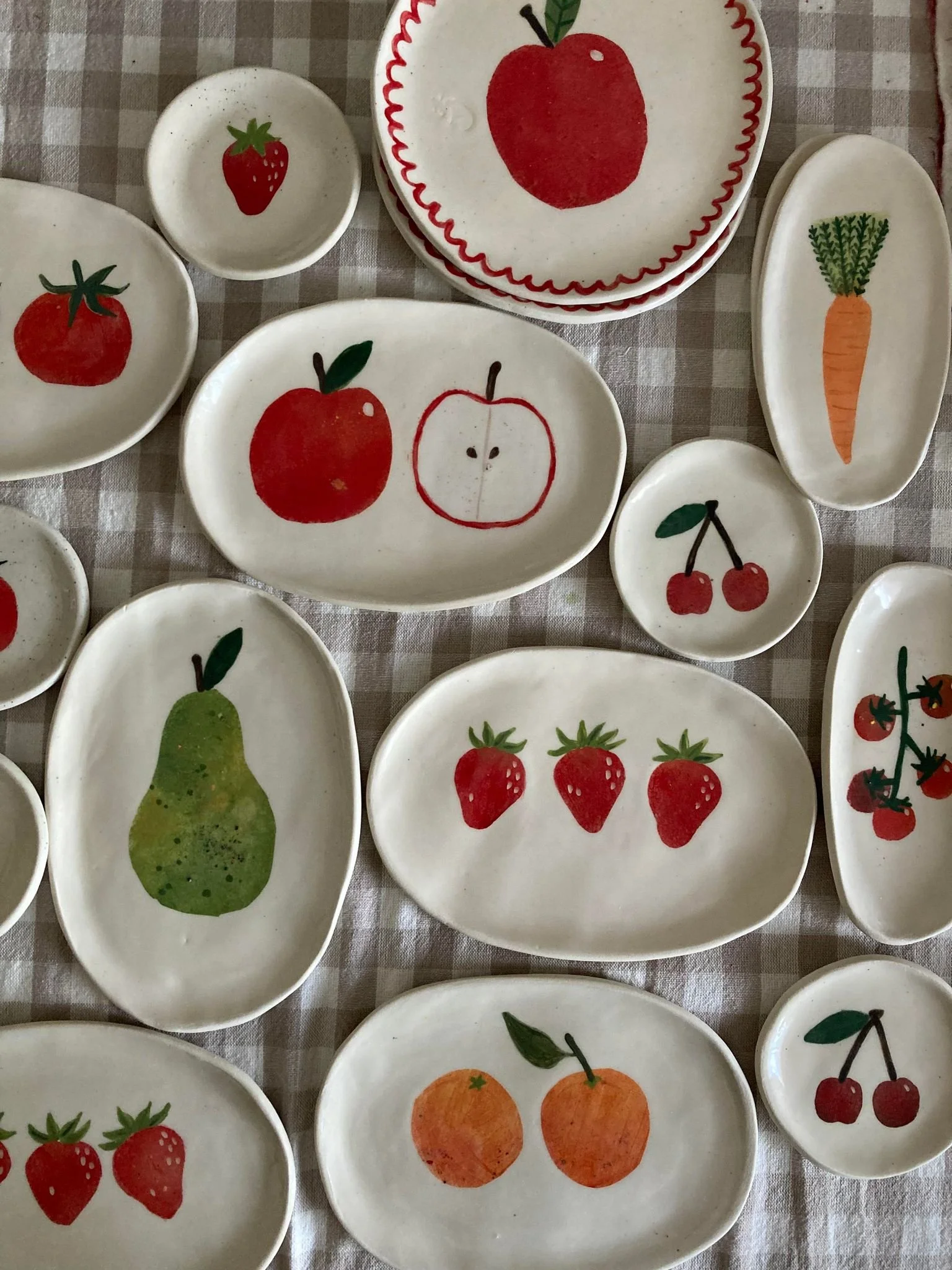 Collection of small decorative plates with painted fruit and vegetable designs, including strawberries, apples, oranges, pears, cherries, carrots, and tomatoes, arranged on a checkered tablecloth.