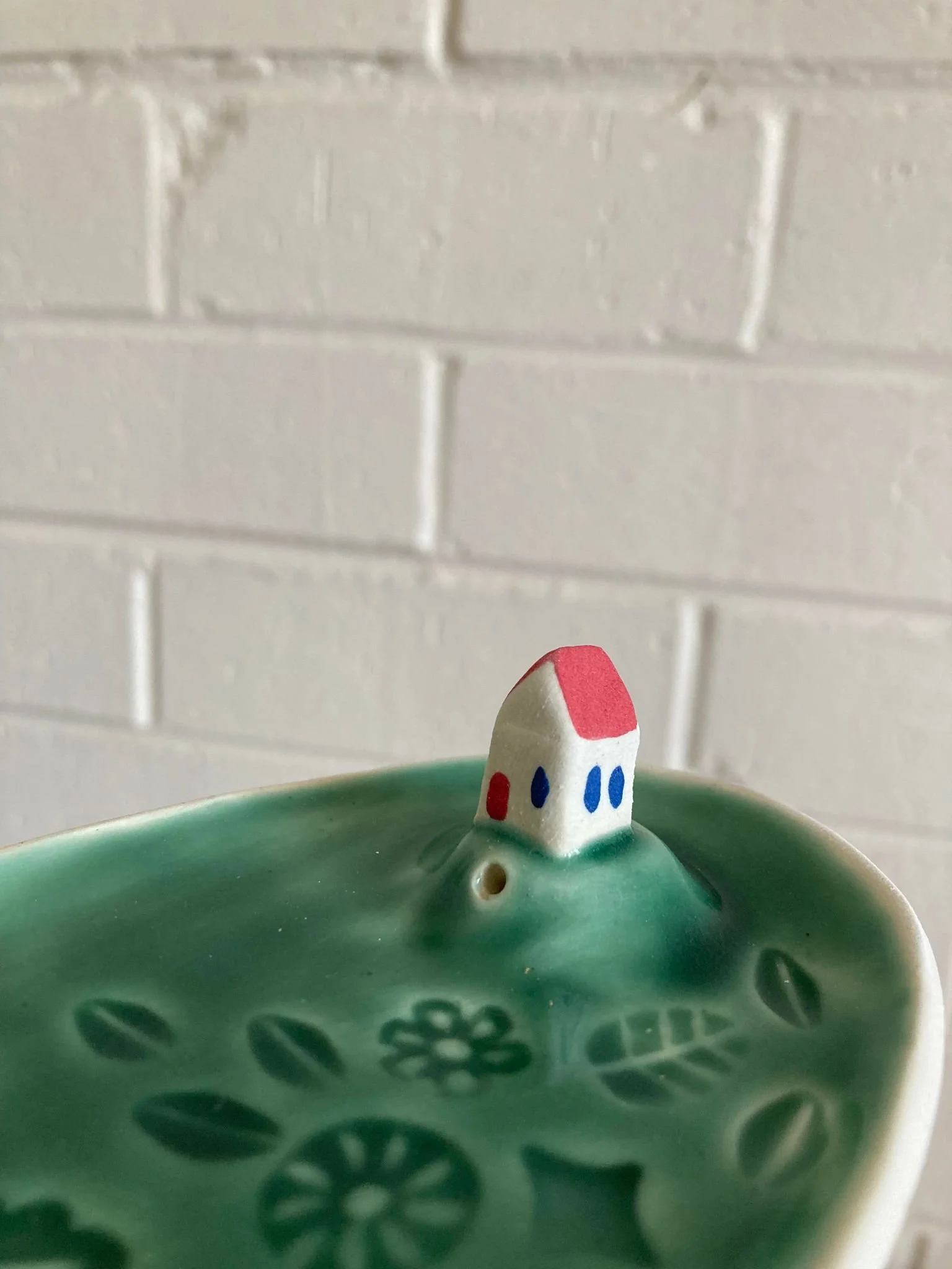Close-up of a decorative ceramic dish with a small house-shaped figurine on its edge, against a white brick wall background.