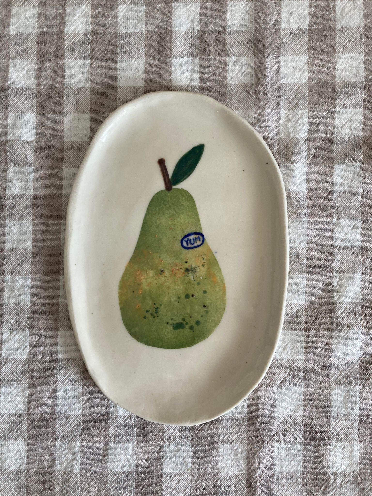 Decorative oval ceramic plate with a painted pear illustration, appraised with the word 'YUM' on a blue sticker, resting on a beige and white checkered fabric surface.