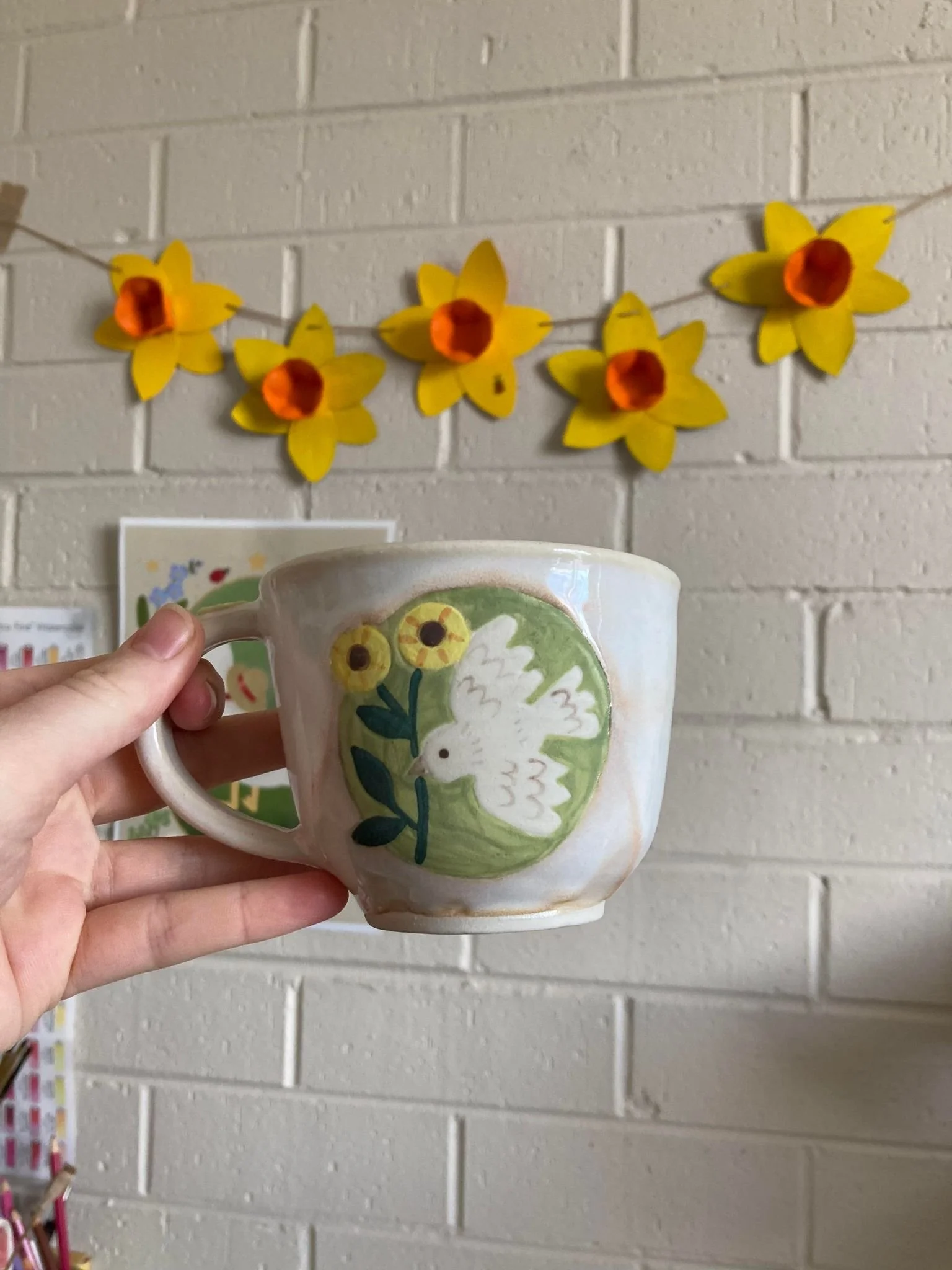 A hand holding a ceramic mug with painted design of a white dove on a green background, surrounded by yellow flowers, against a brick wall decorated with yellow paper daffodil garland.