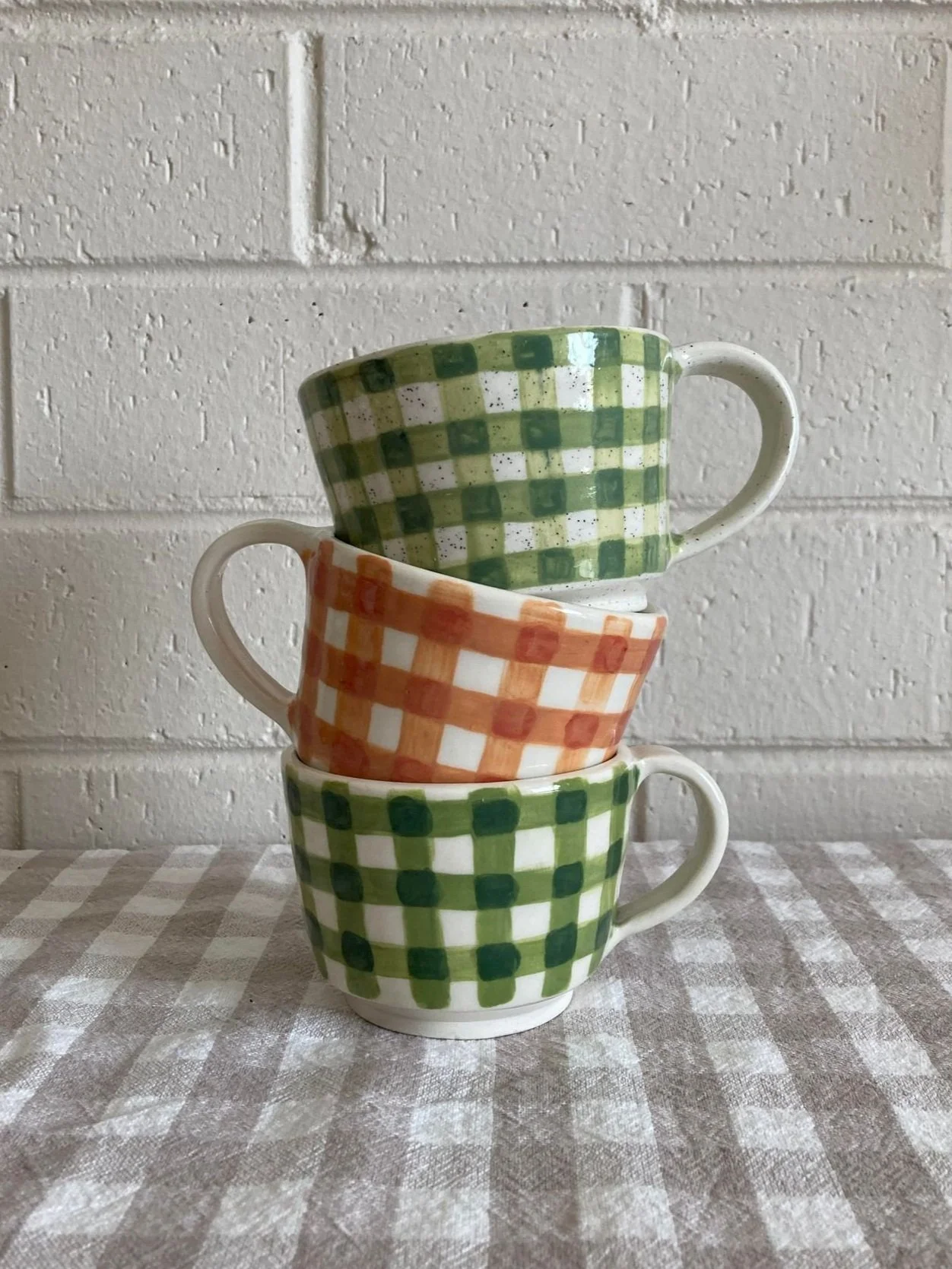 Stack of three ceramic cups with green and orange checkered patterns, placed on a checkered tablecloth against a white brick wall.