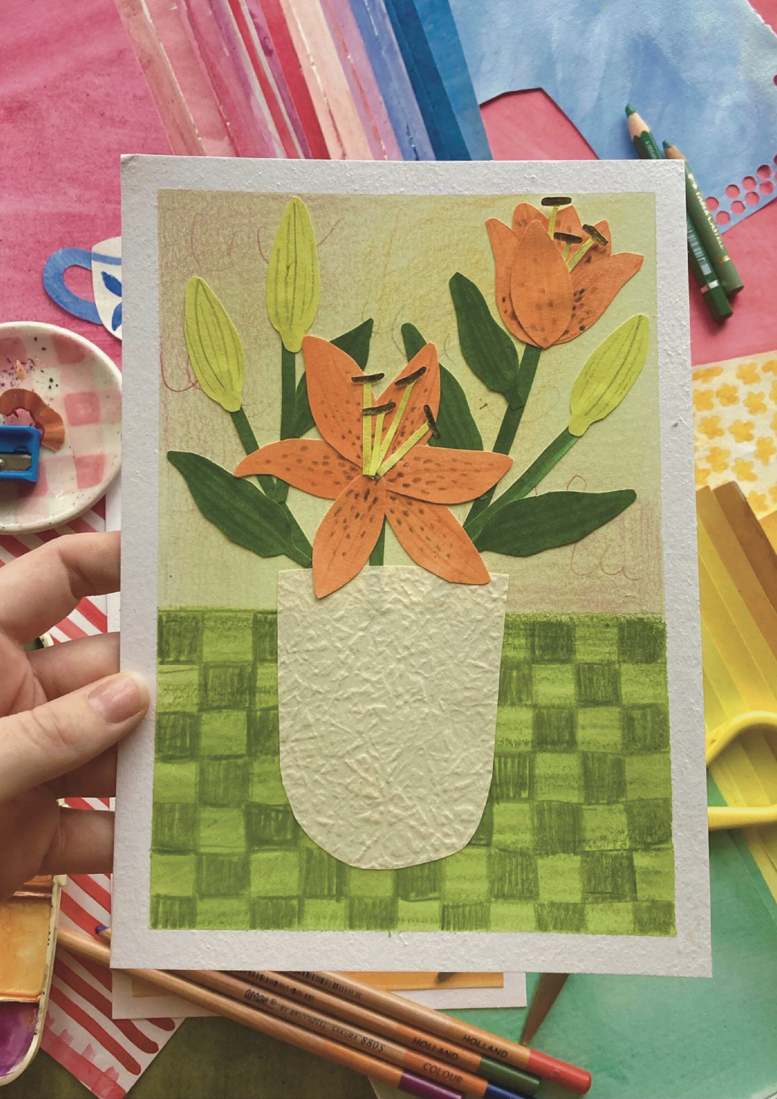 Handmade greeting card featuring an orange lily flower in a white textured pot, with green leaves and buds, placed on a green checkered tablecloth.