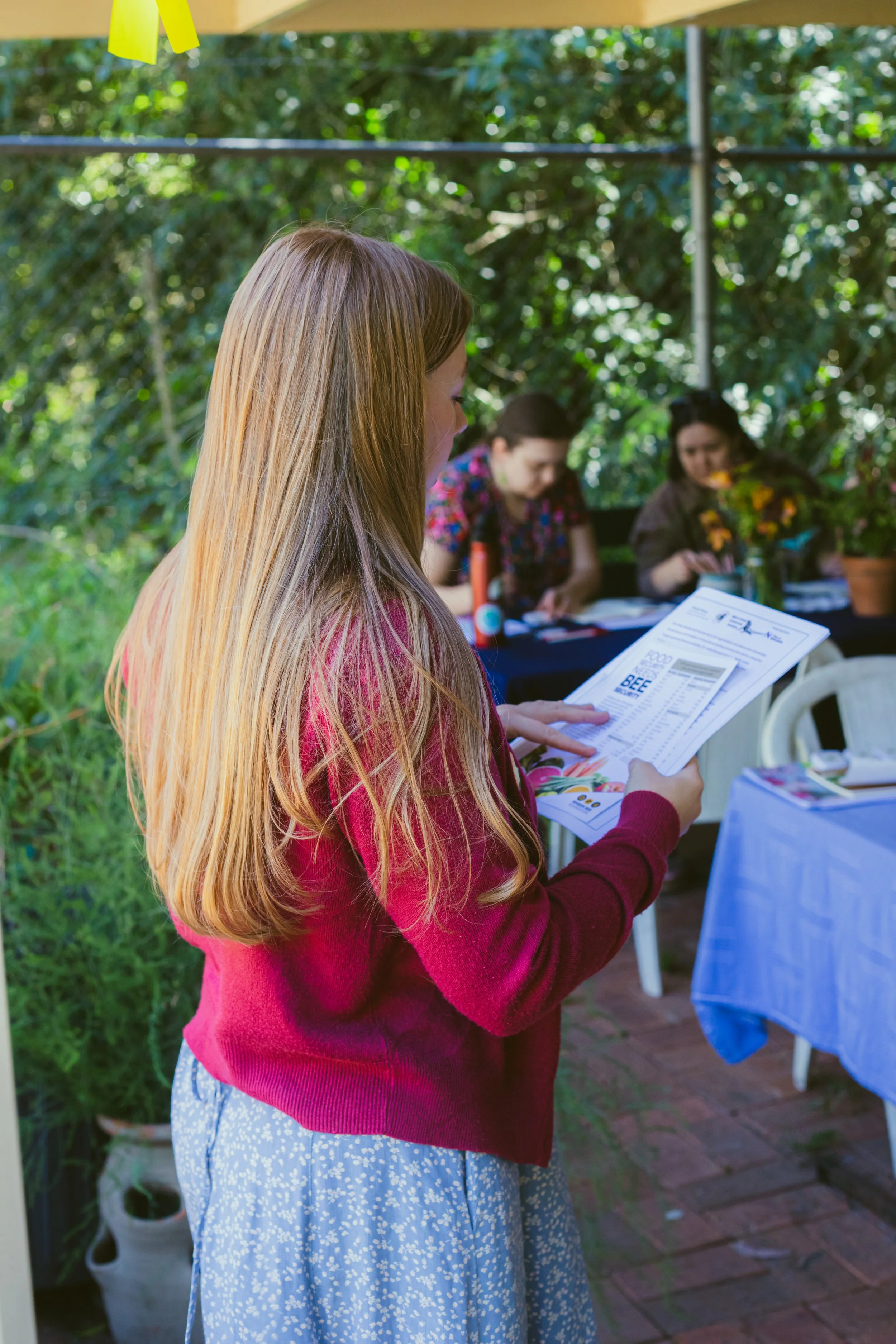 Gemma with long ginger hair reading a document outdoors at a gathering, with two women seated at a table in the background.