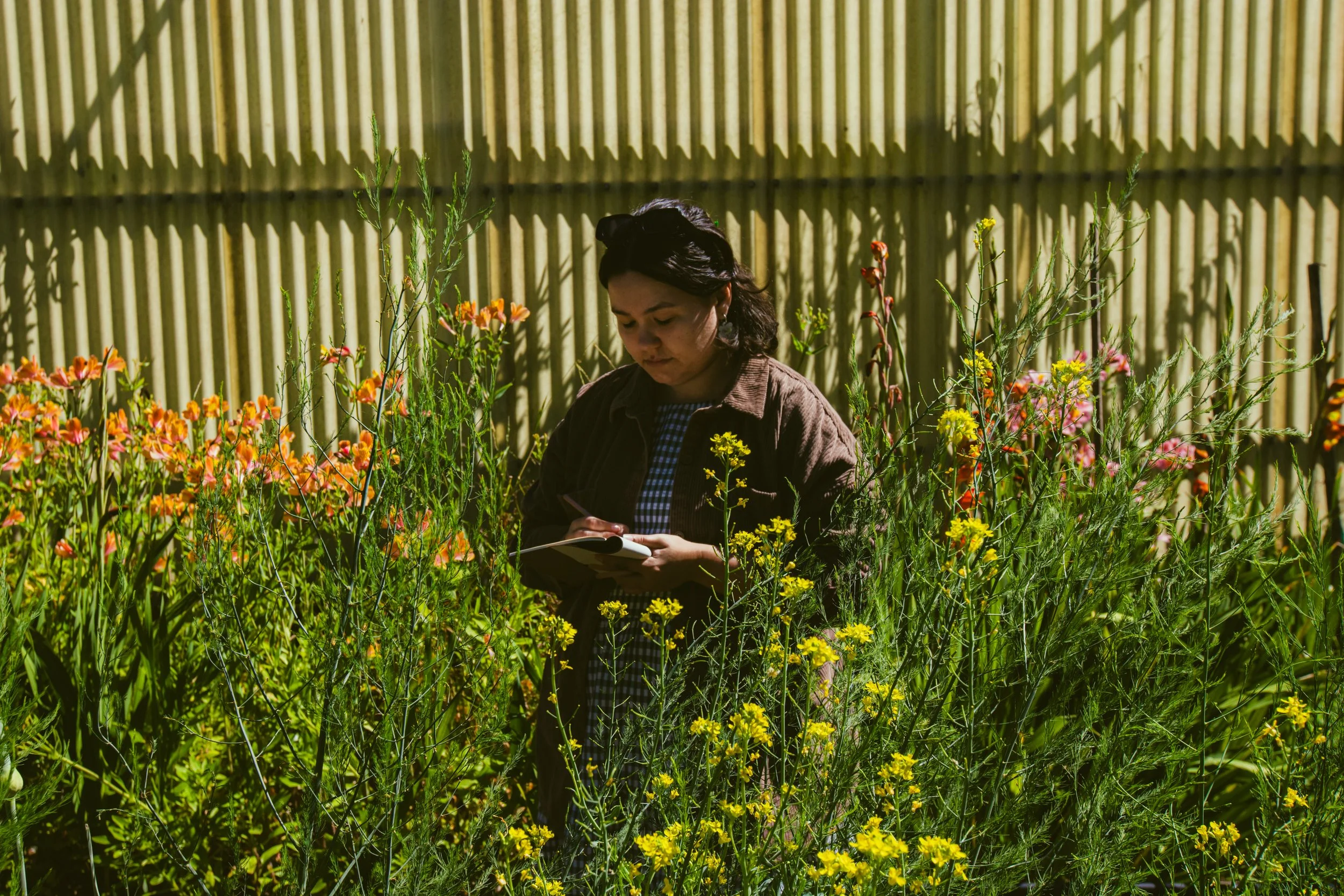 A woman standing in a garden with yellow and pink flowers, writing in a notebook.